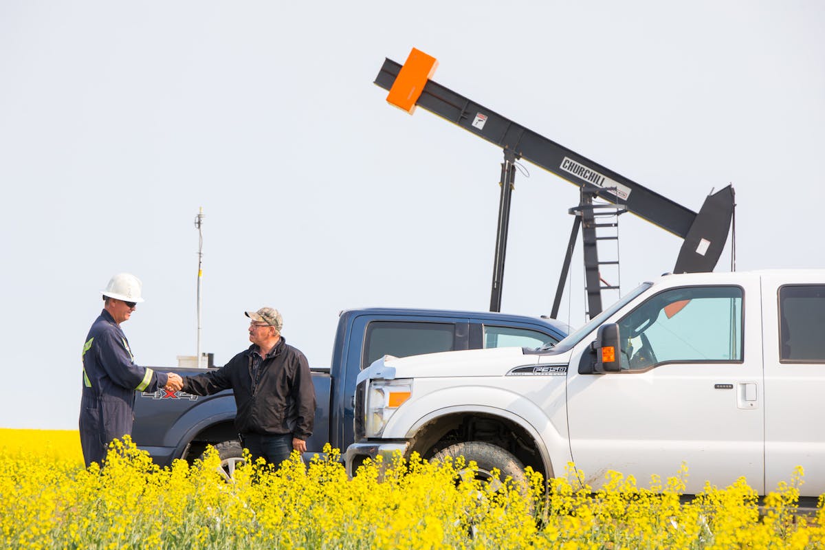 Two men shaking hands in a canola field, in front of oil machinery.