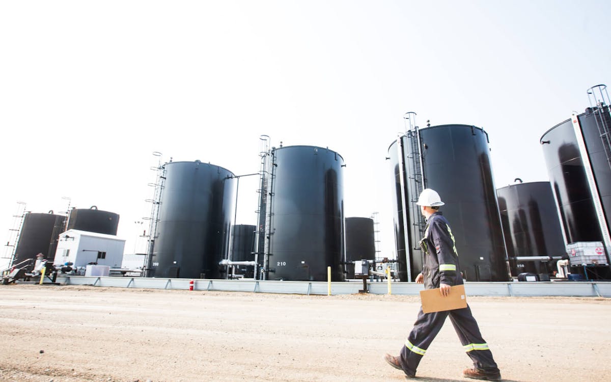 A worker in a dark uniform, walking in front of large vats on an oil rig.