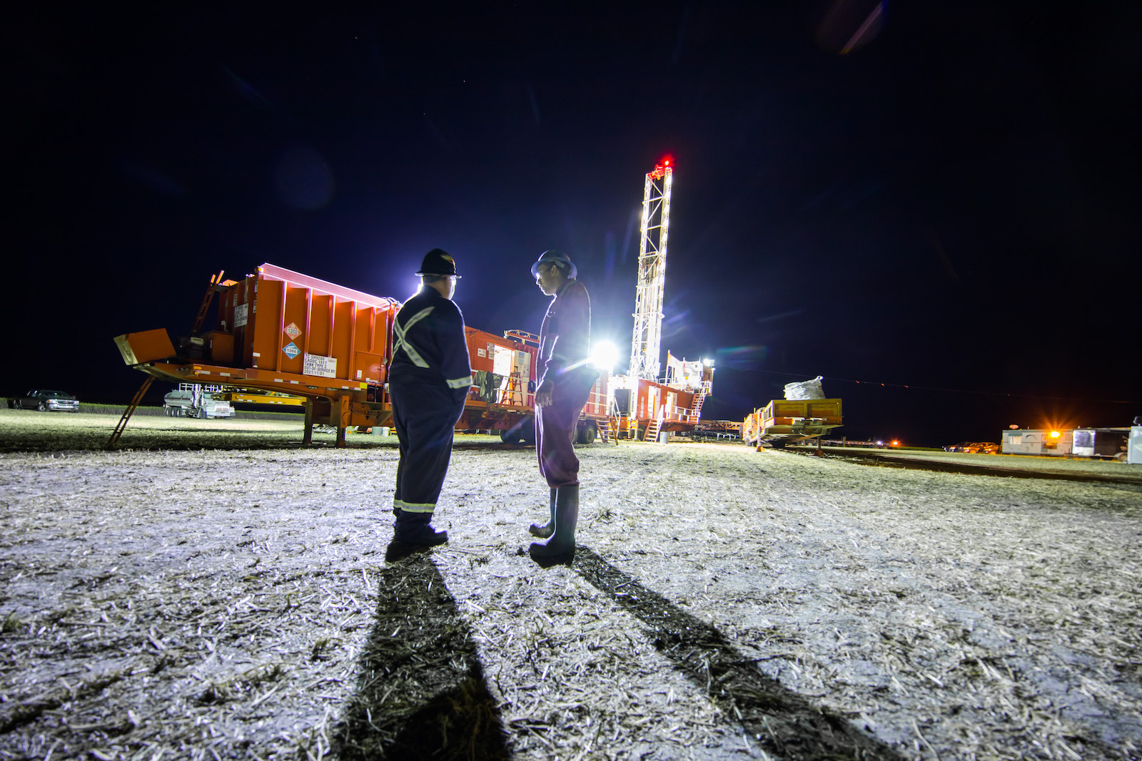 Two oil rig workers chat near an oil rig at night, backlit by a bright light.