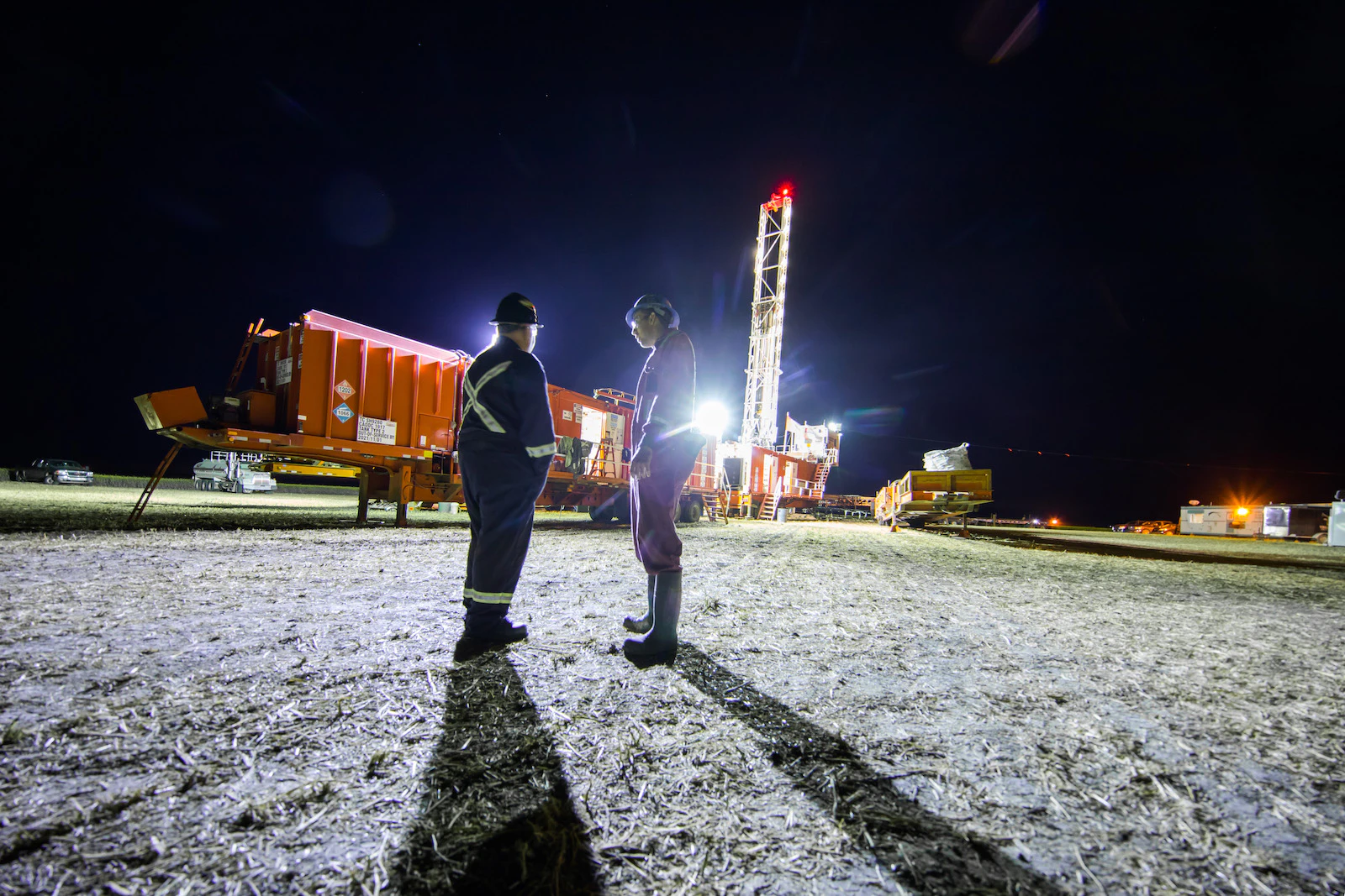 Two oil rig workers chat near an oil rig at night, backlit by a bright light.