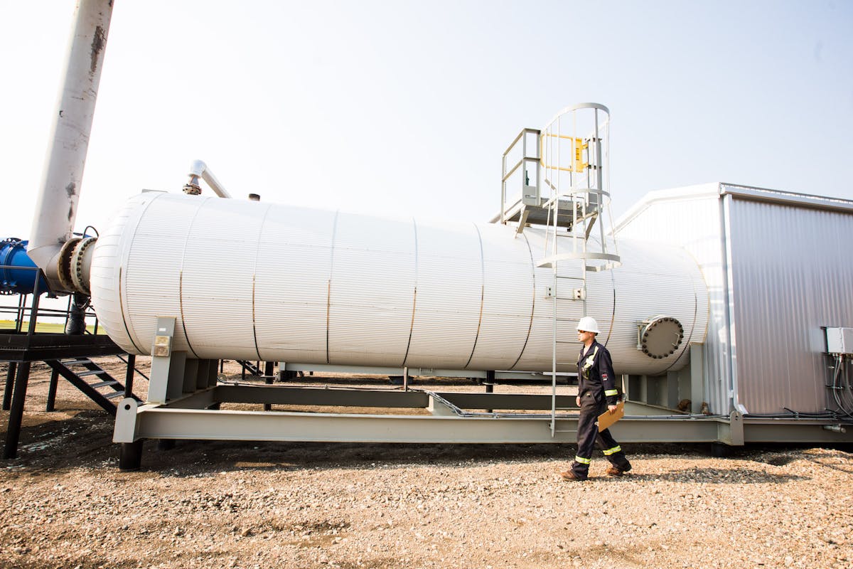An oil worker walking with a clipboard, in front of a large white tank.