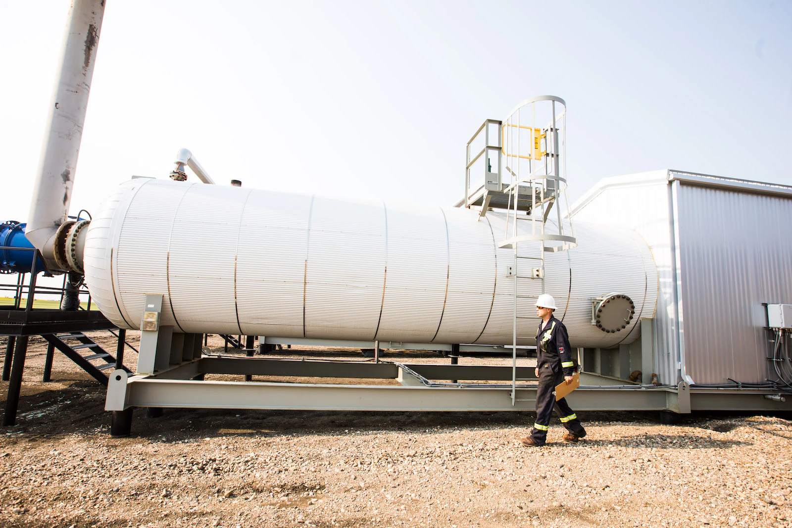 An oil worker walking with a clipboard, in front of a large white tank.