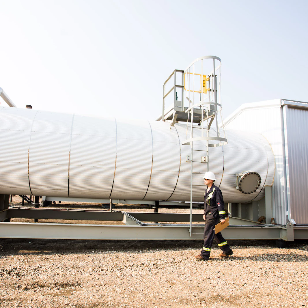 An oil worker walking with a clipboard, in front of a large white tank.