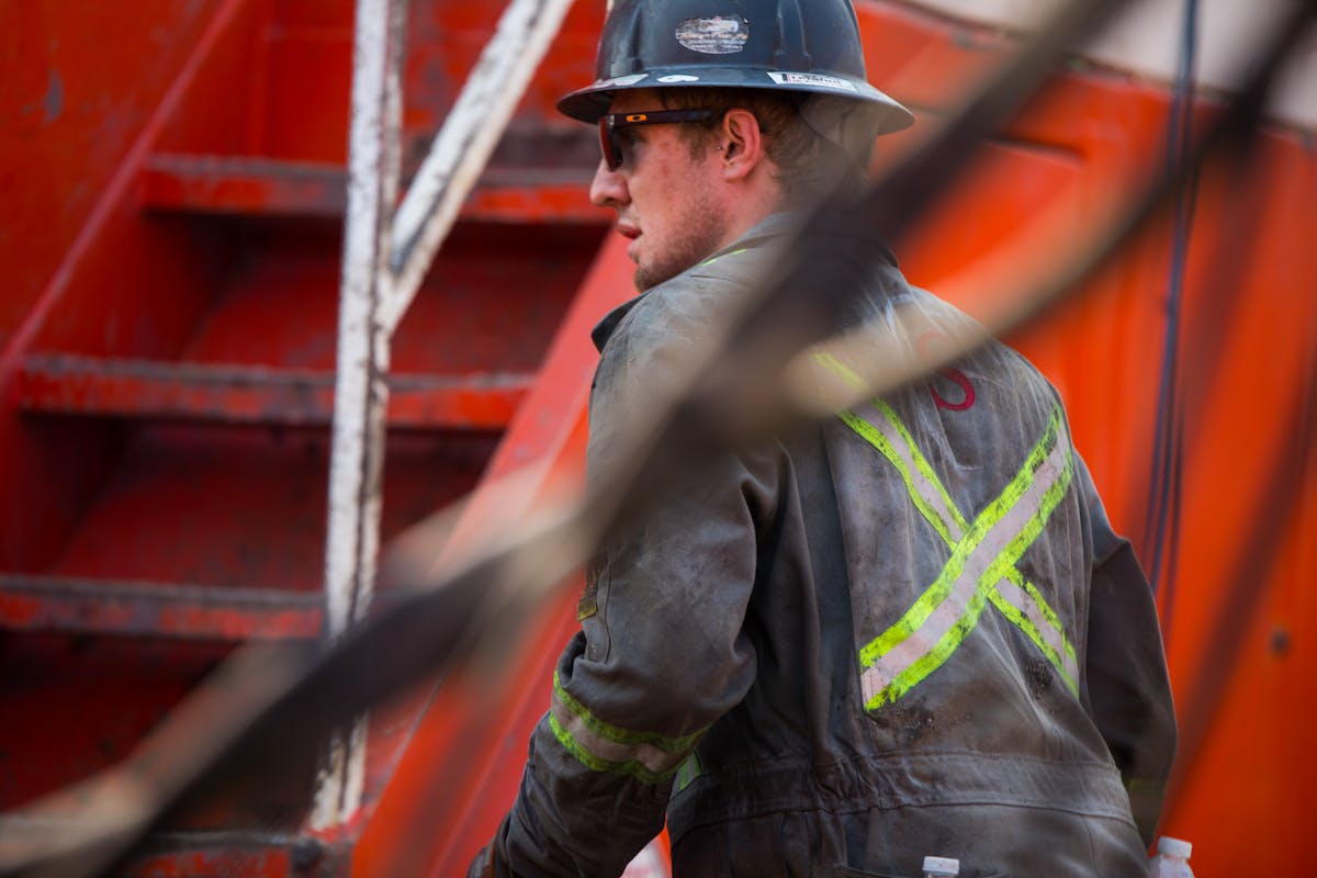 An oil rig worker wearing a safety jacket and hard hat walks next to a red ladder.