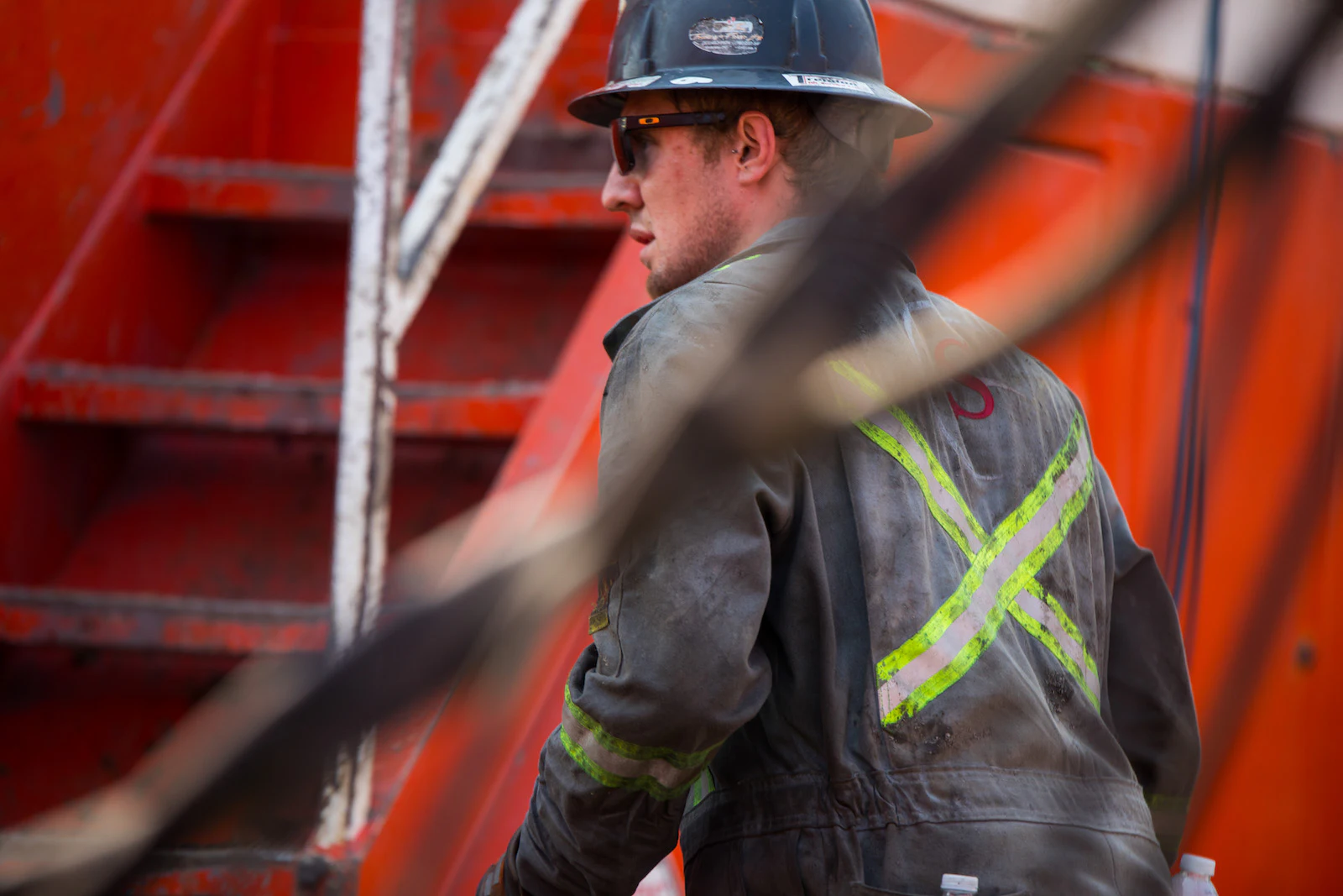 An oil rig worker wearing a safety jacket and hard hat walks next to a red ladder.
