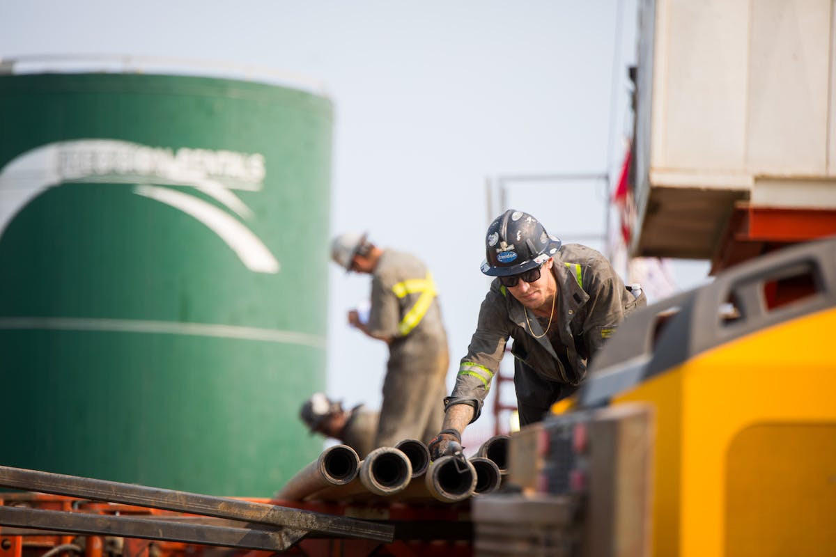 Two oil workers move pipes near a green oil drum.