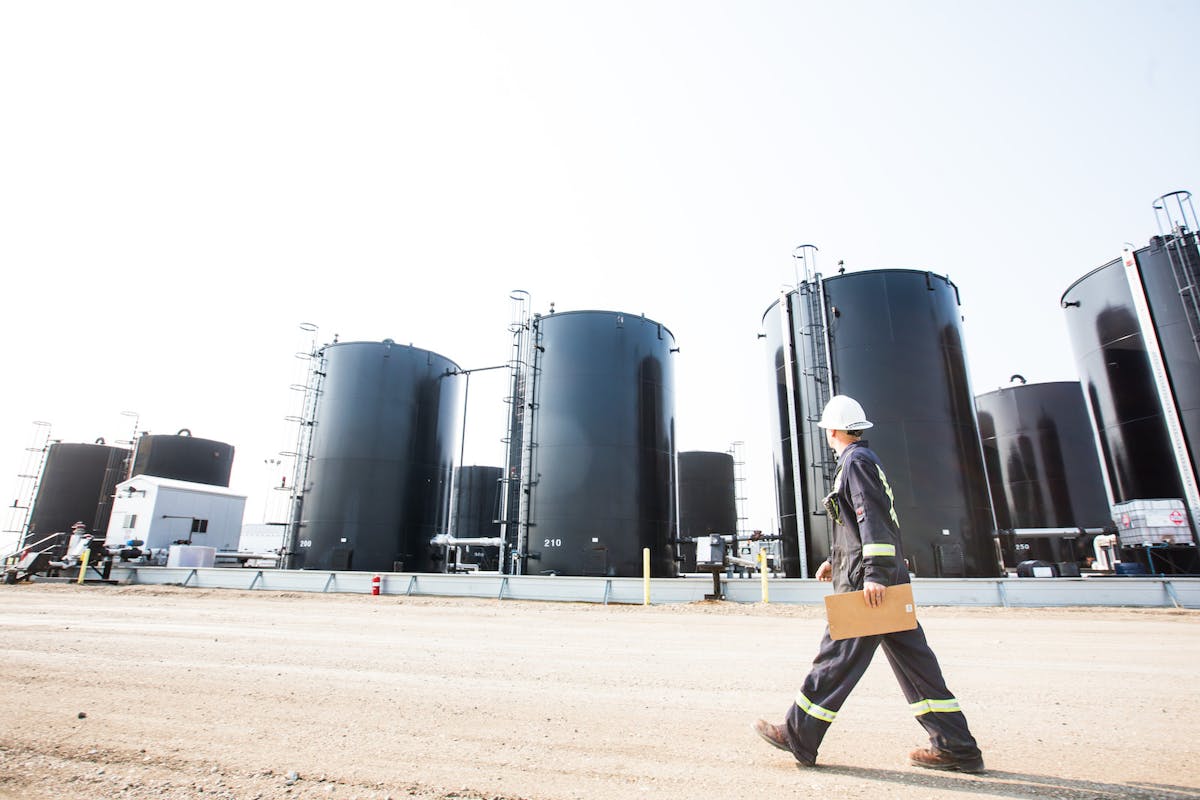 An oil rig worker, walking with a wide-legged gait in front of several large oil drums.