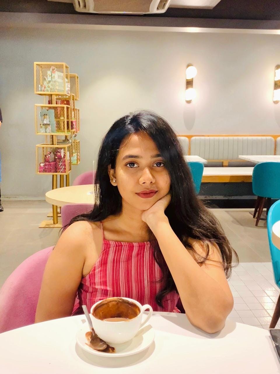 A woman sitting in a cafe with a cup of hot chocolate, wearing a red sleeveless top and resting her chin on her hand.