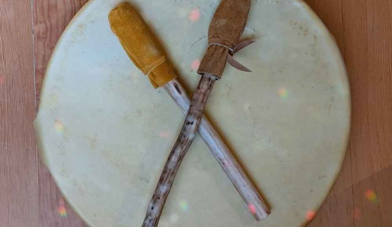 A 10" round indigenous animal hide drum with two drumsticks laying on top of the drum. 