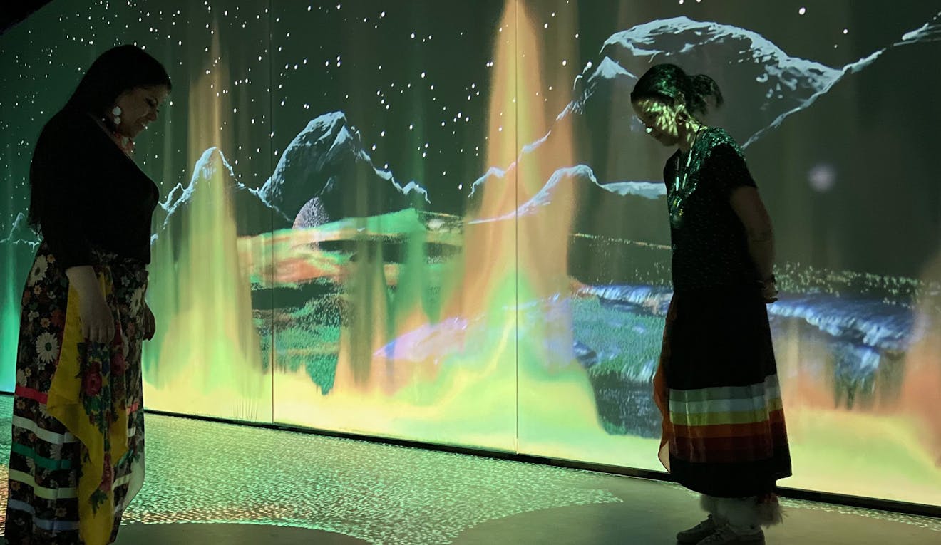 Two women wearing colourful ribbon skirts are in an immersive pixel playground with the colours of the northern lights surrounding them from floor to ceiling.