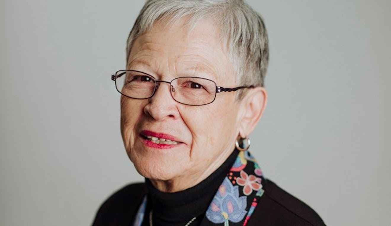 An older indigenous lady with grey hair wearing glasses and red lipstick sits for her headshot picture.