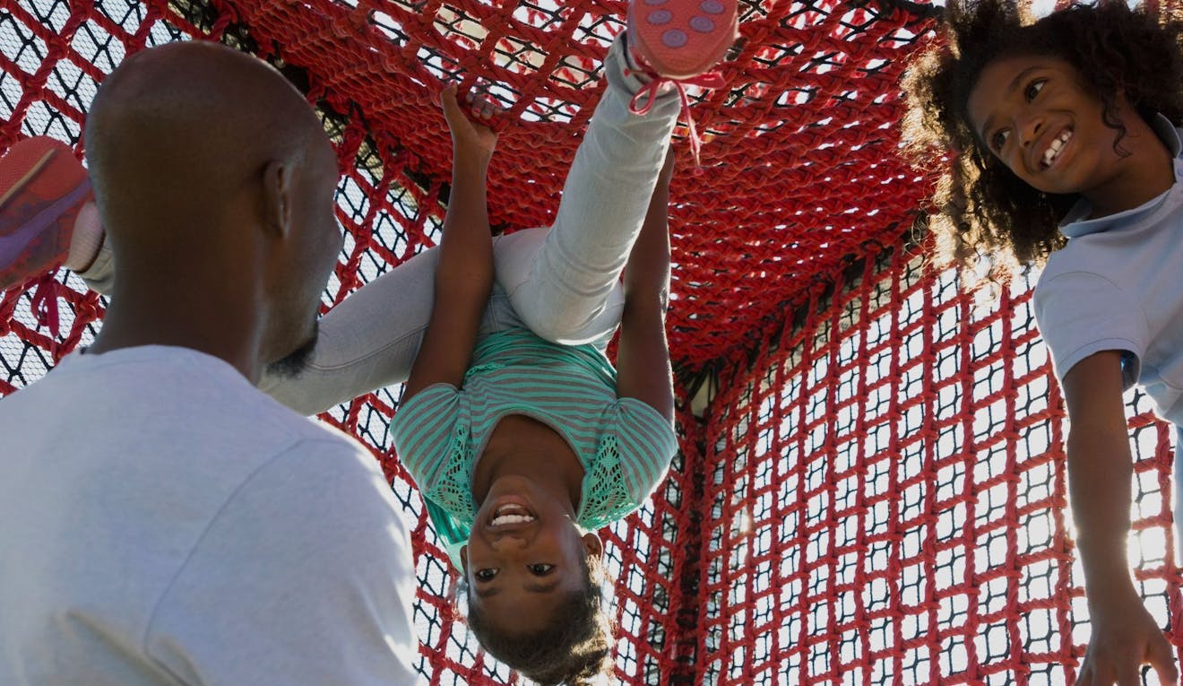 A father climbs a netted tower with his two daughters in a playground. One hangs upside down, the other swings with one arm holding on.