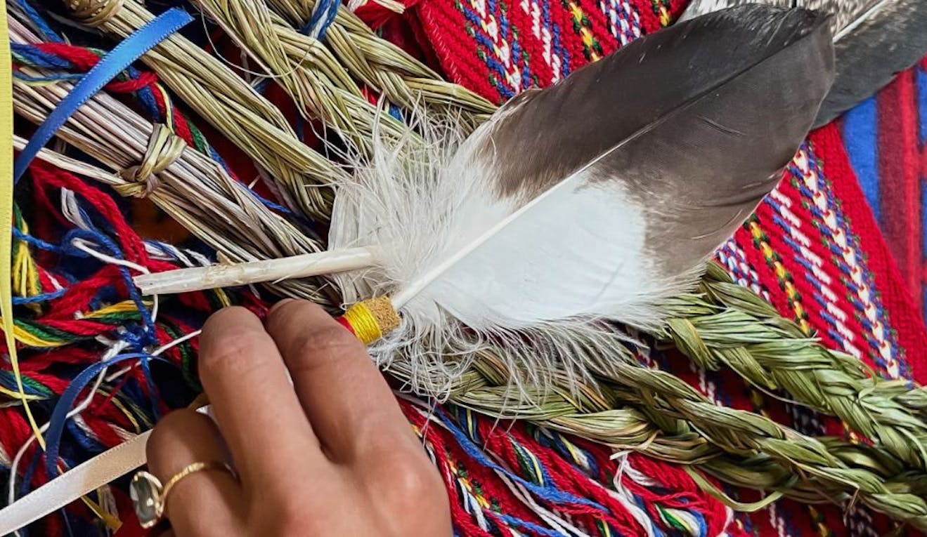 A hand places a feather on top of braided sweetgrass and a metis scarf.