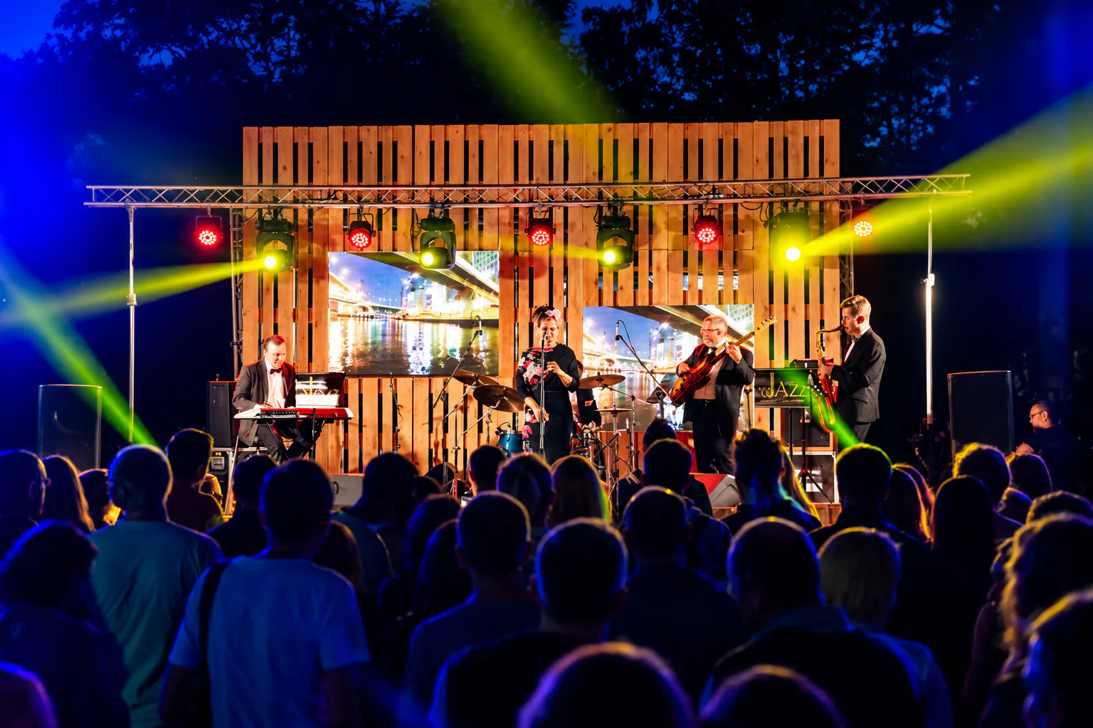 Jazz band playing on a wooden pallet stage with colored lights for an evening crowd