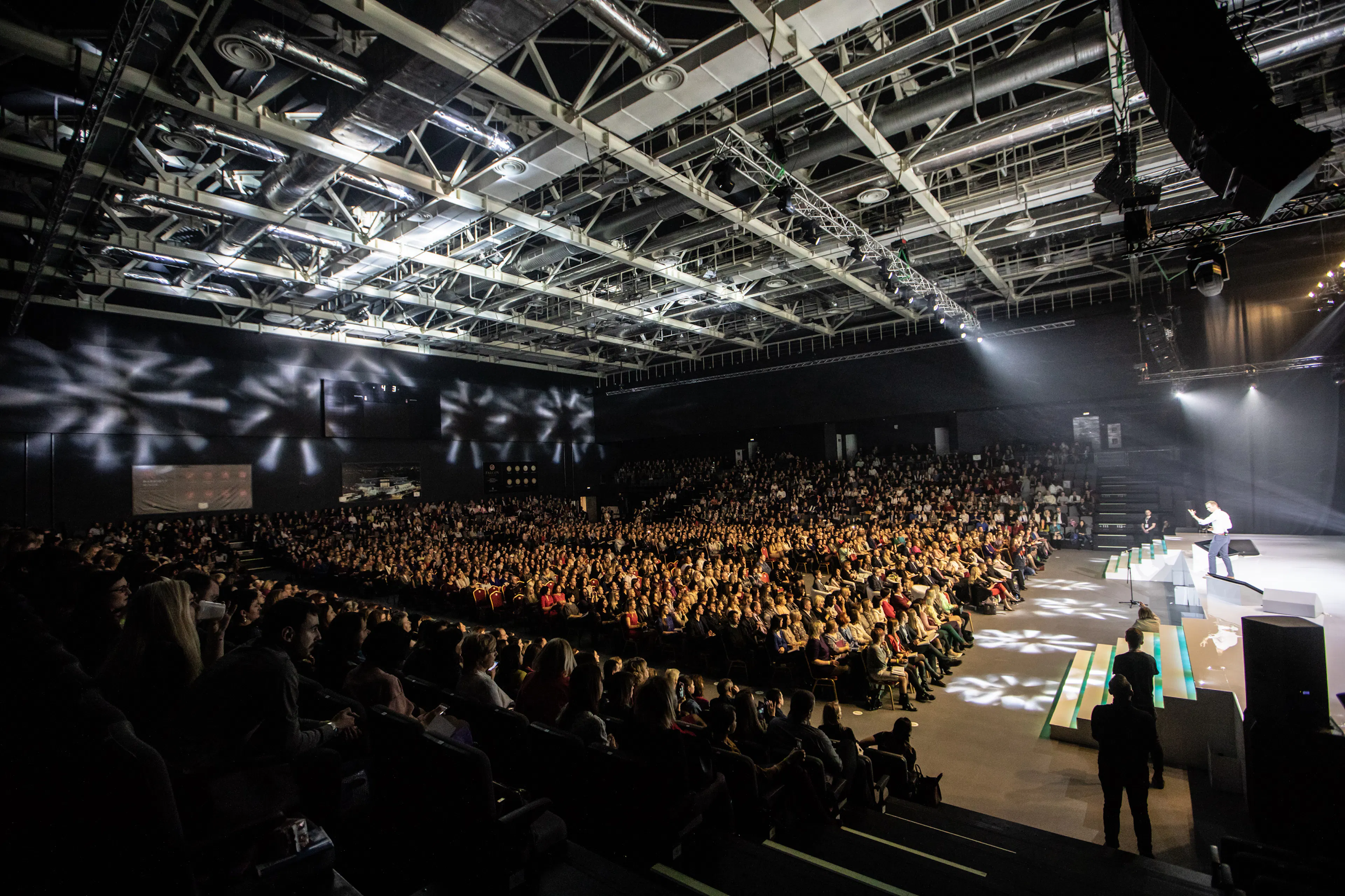 Szeroki widok sali konferencyjnej z prelegentem na oświetlonej scenie i pełną widownią Summit Oriflame