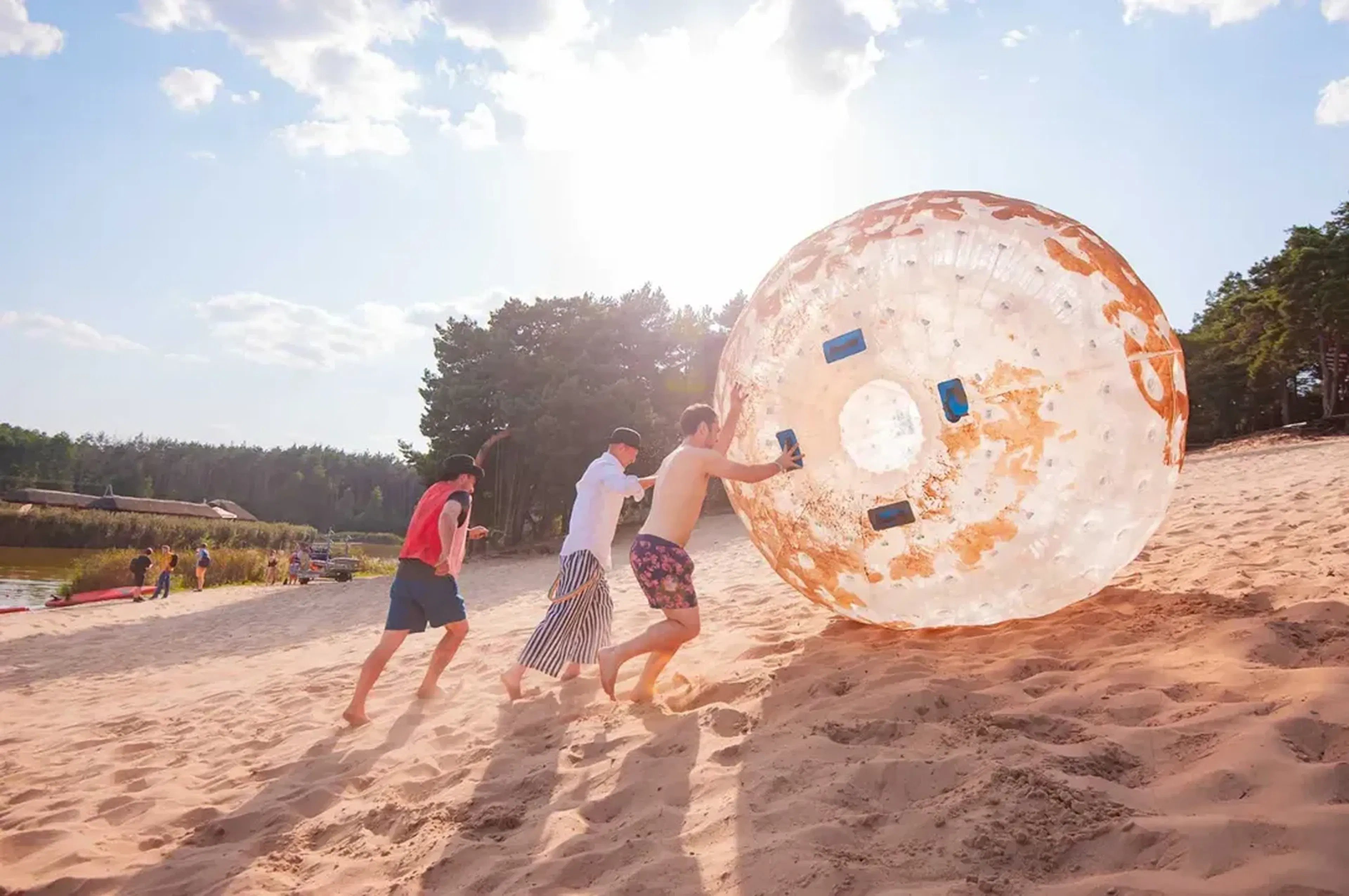 Pracownicy Godel pchają ogromną dmuchaną kulę zorb po słonecznej, piaszczystej plaży nad jeziorem