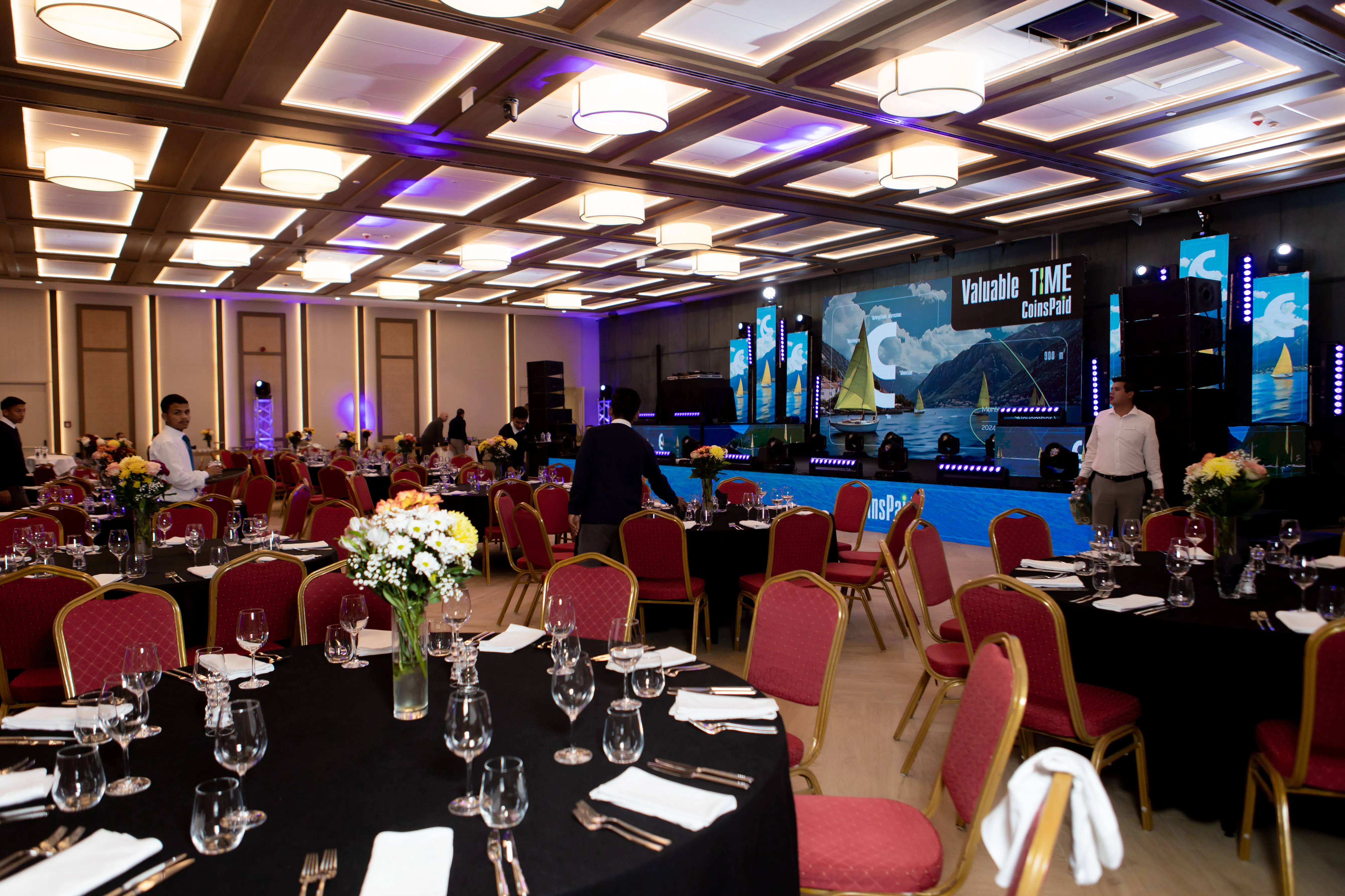 Banquet hall set with round tables and red chairs facing the CoinsPaid Valuable Time LED stage