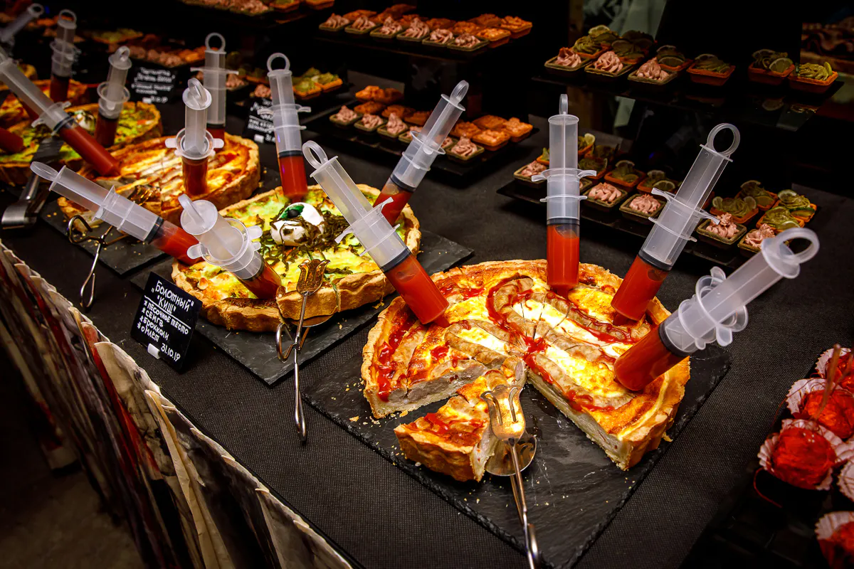 Halloween buffet of quiches pierced with syringes of red sauce, surrounded by themed canapés