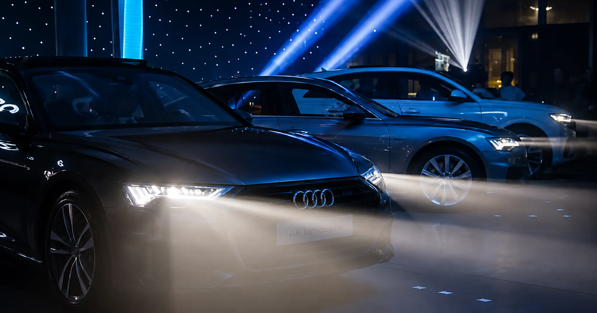 Audi A6 Limousine with headlights on, lined up with other Audi models under blue light beams and a starry backdrop