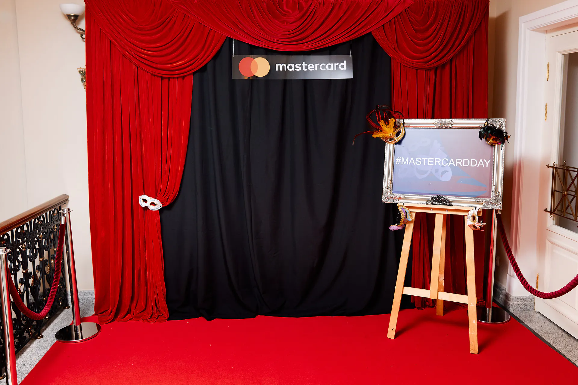 Masterday photo zone with red velvet curtains, a Mastercard sign and a #MASTERCARDDAY easel with carnival masks
