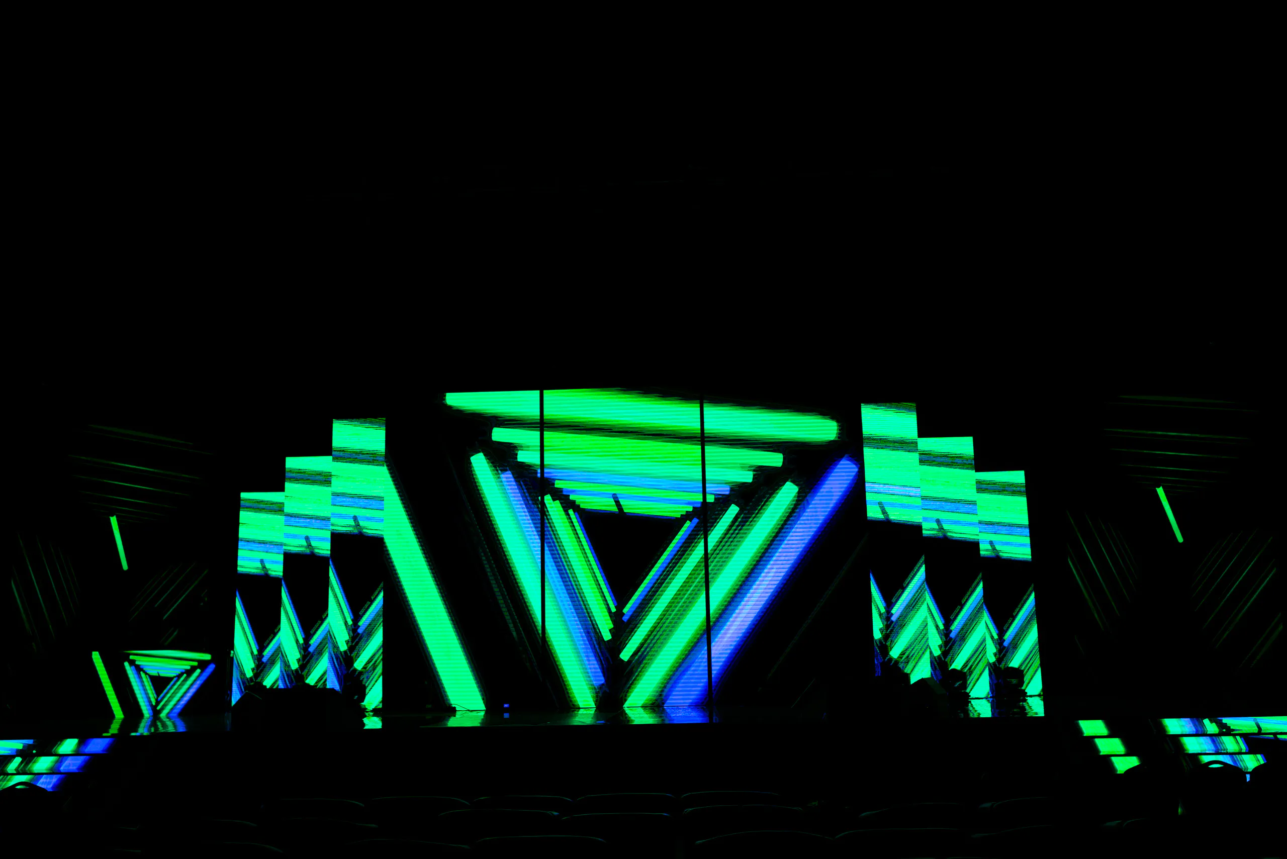 Multimedia stage set with green and blue geometric visuals on LED screens in a darkened hall