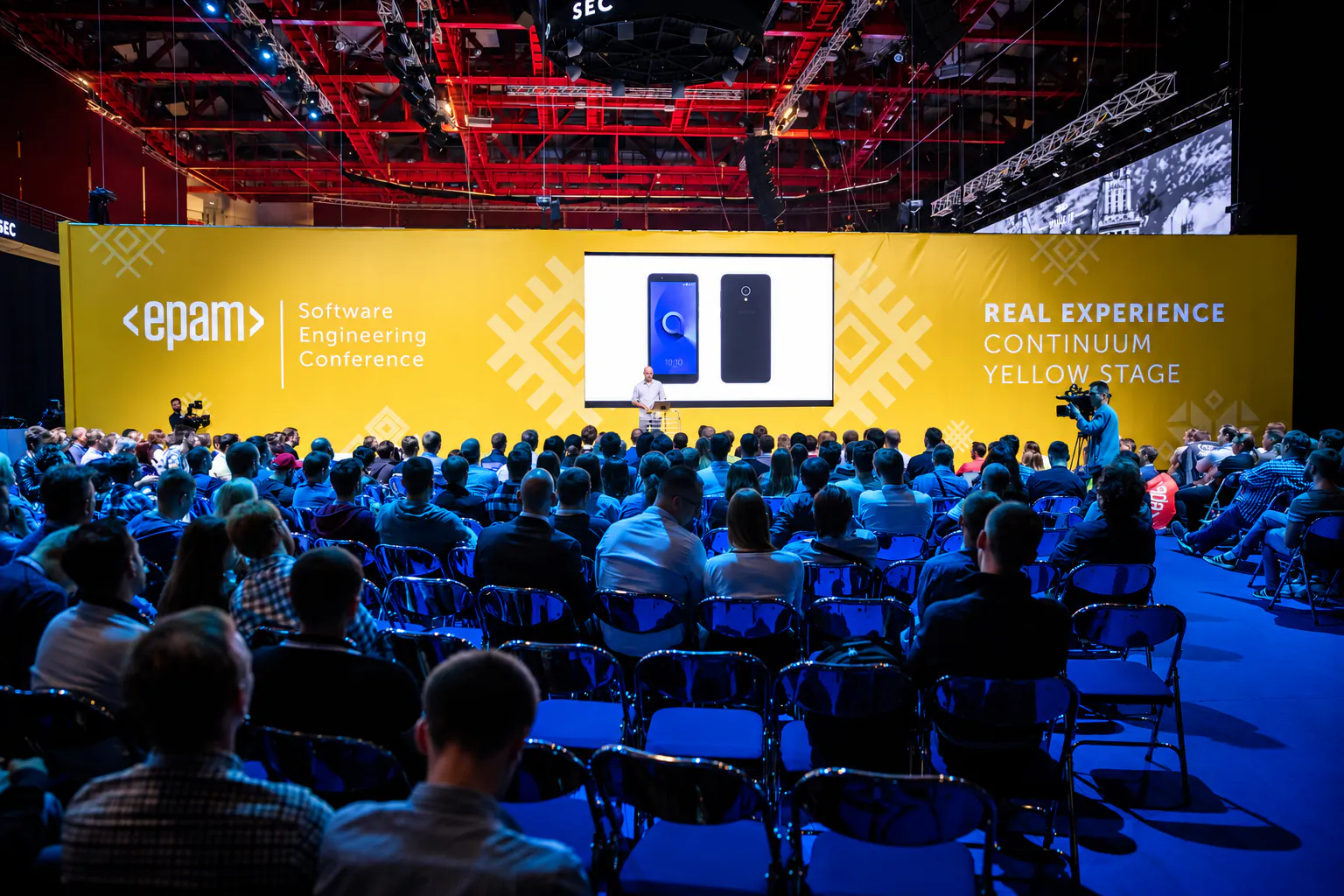 Audience facing a speaker on the EPAM Software Engineering Conference yellow stage under red roof trusses