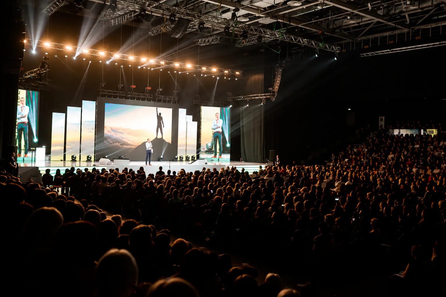 Speaker on a stage flanked by LED screens under a lighting truss, facing a packed Oriflame Summit hall