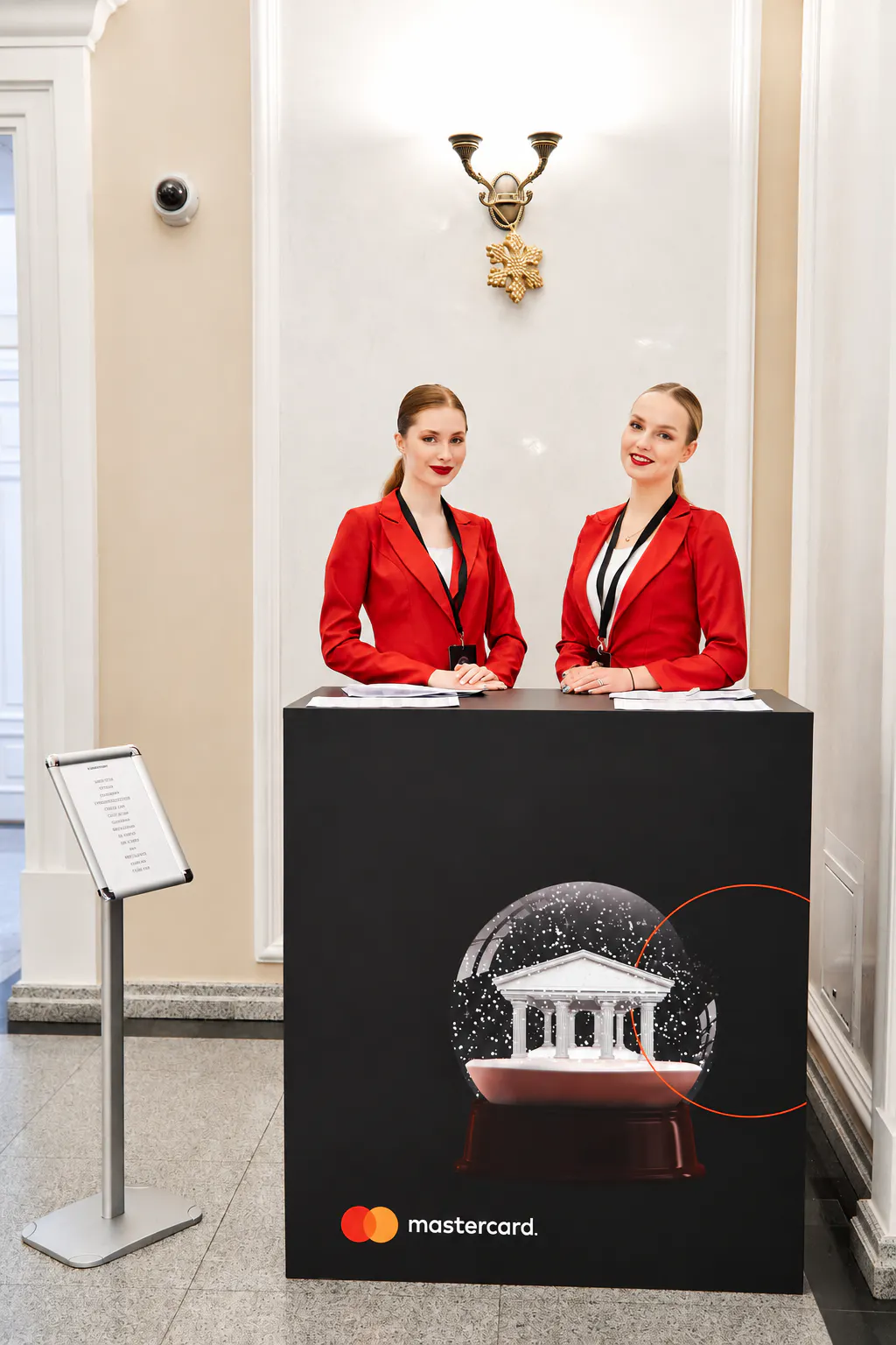 Two hostesses in red blazers at a Mastercard-branded welcome desk with a snow globe graphic