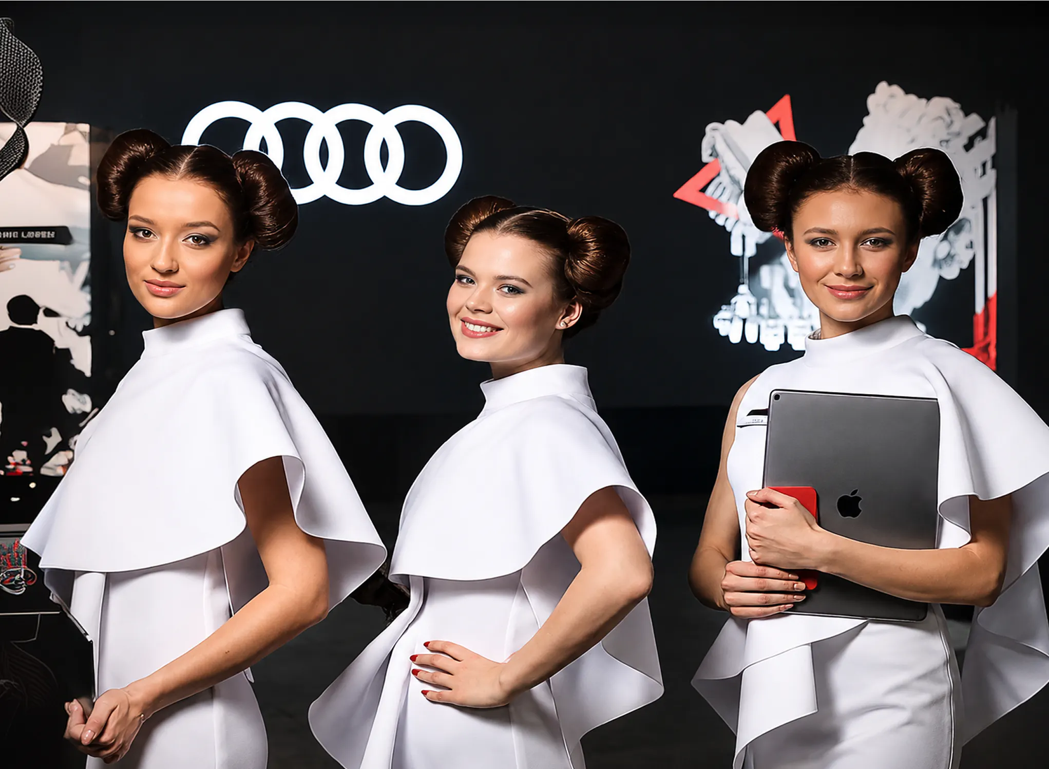 Three hostesses in white caped dresses posing in front of the illuminated Audi rings logo