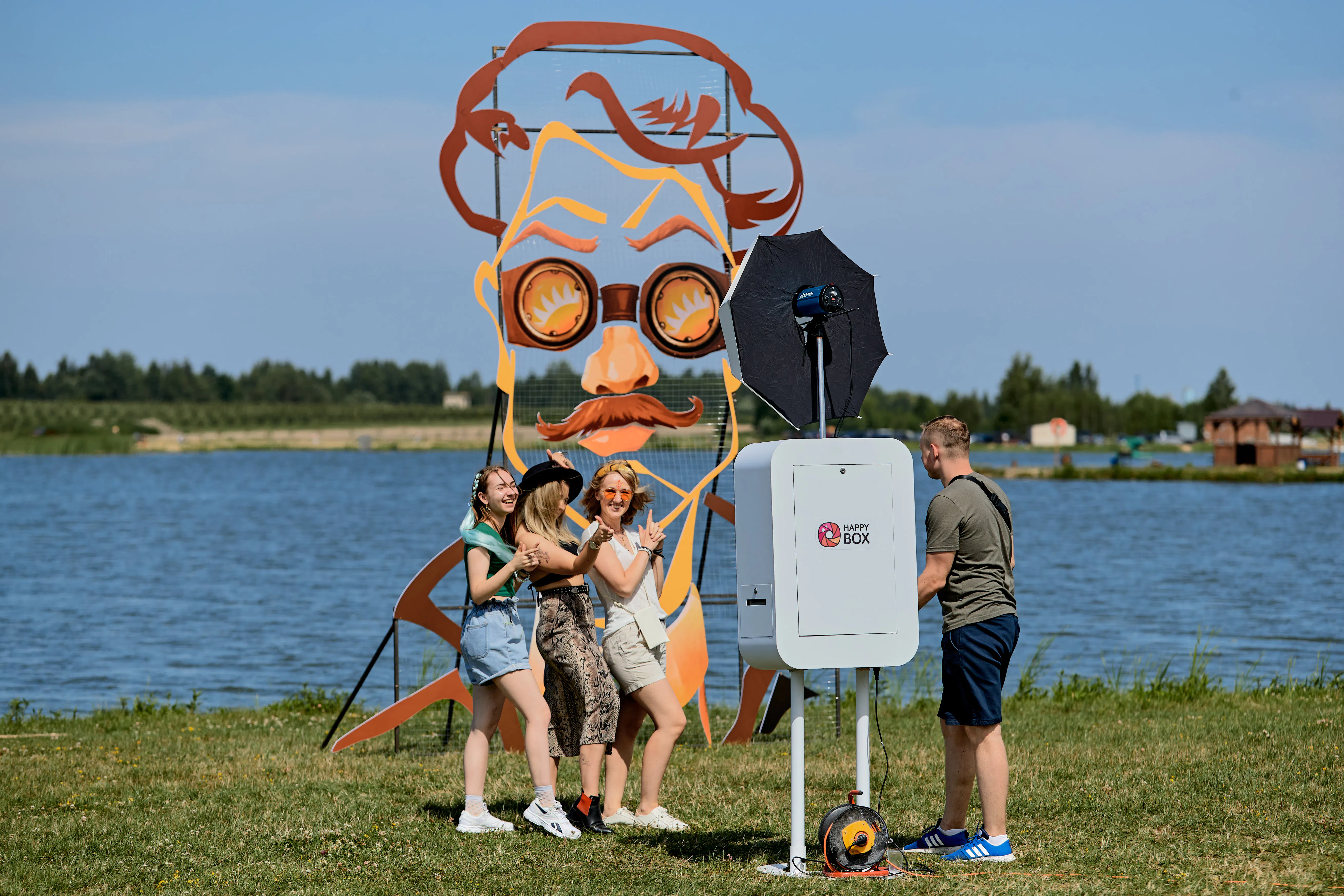 Guests posing at a Happy Box photo booth next to a giant mustached-face art installation by the lake