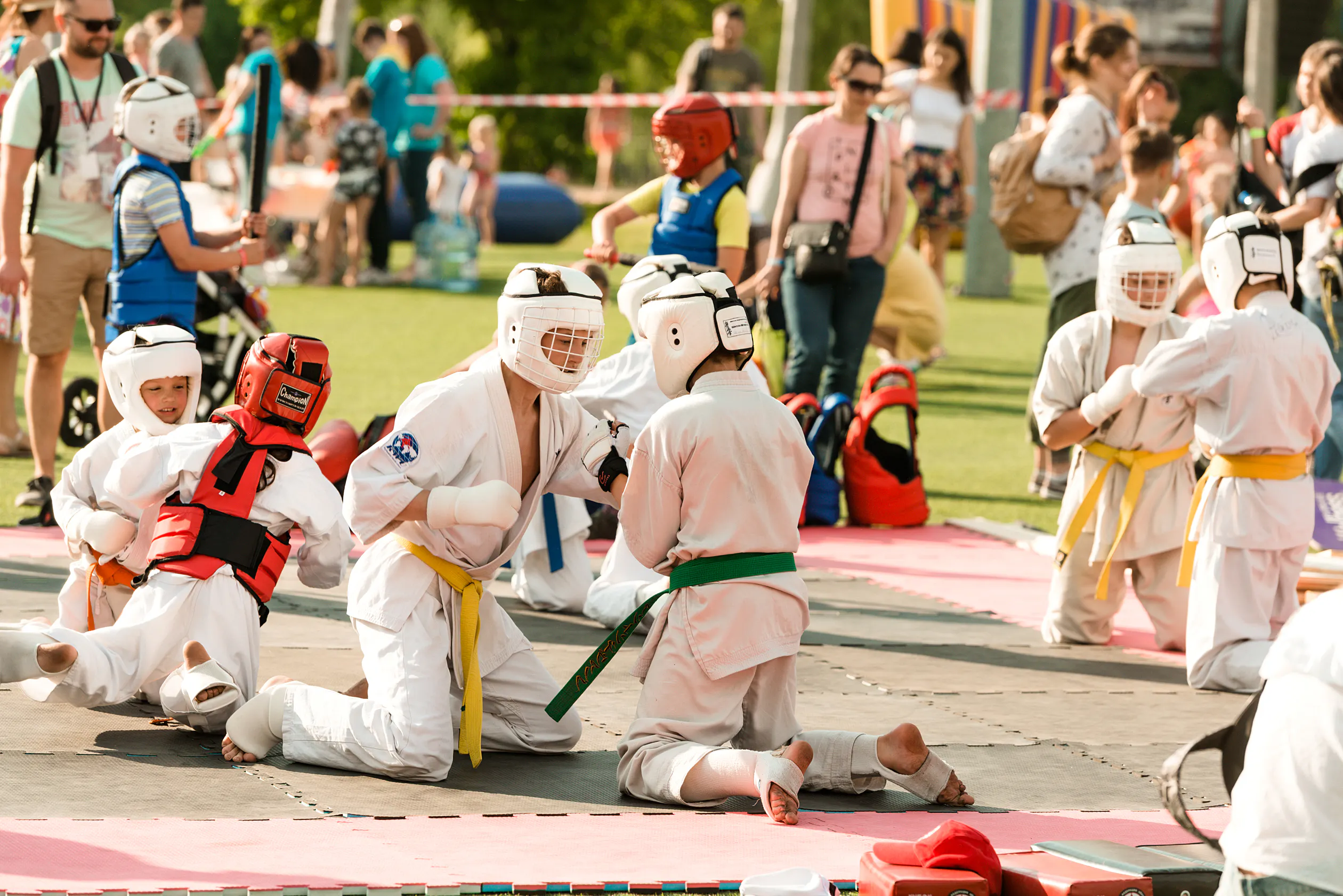 Kids in karate uniforms and protective headgear sparring on mats in the park