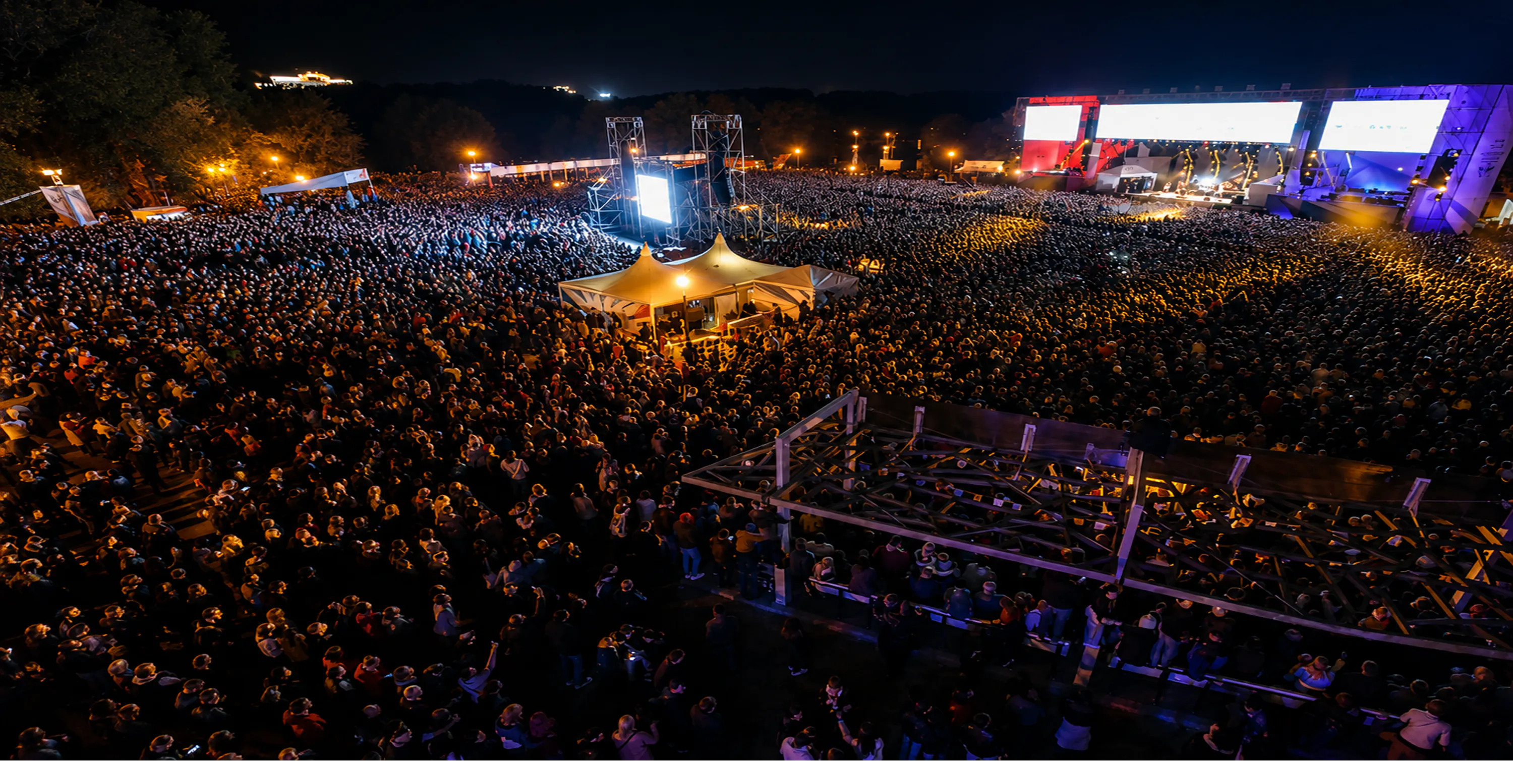 Night view over the packed Wargaming Fest grounds with two lit stages, LED screens and a central tent