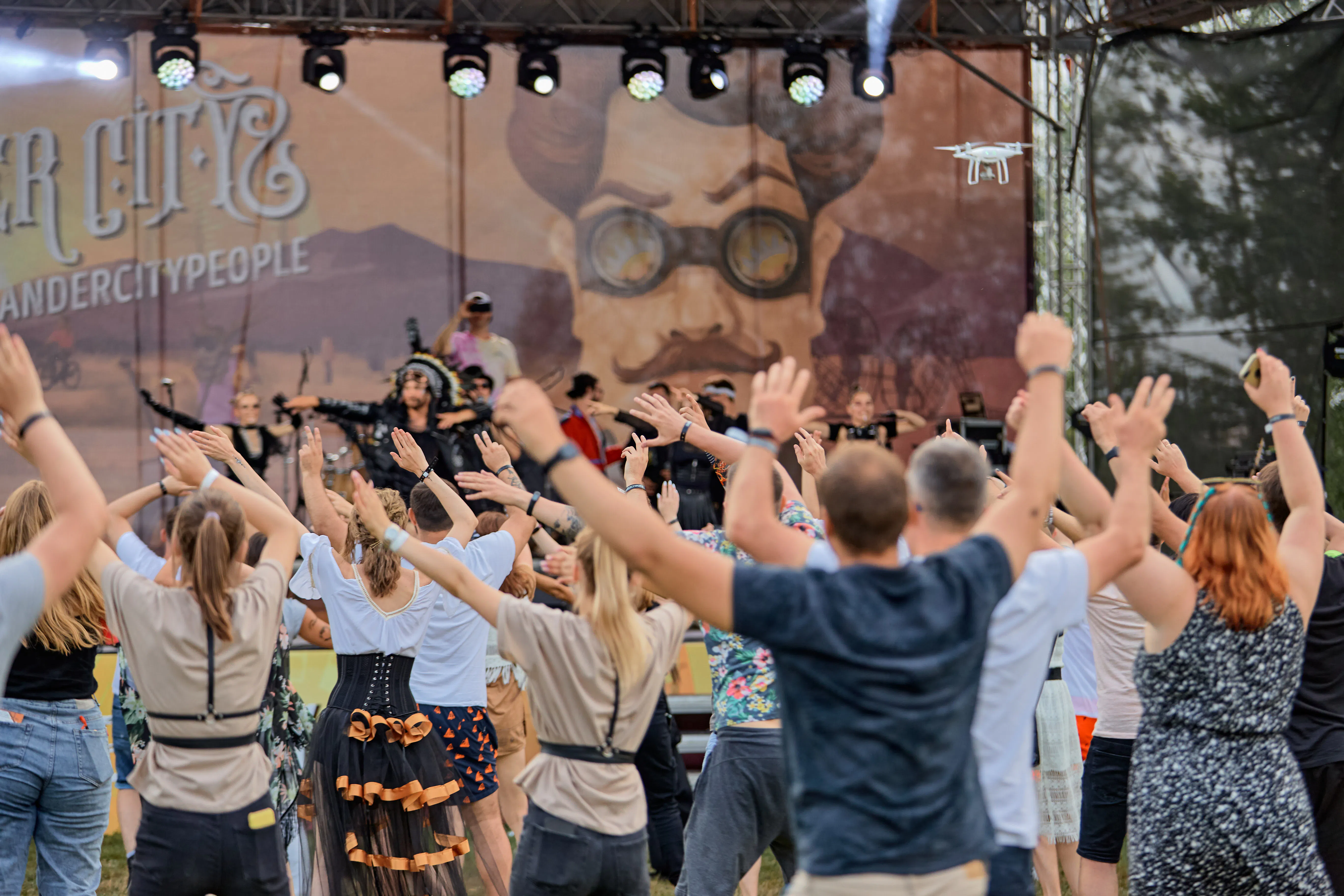 Costumed performers on the Ander City stage leading a crowd with raised hands as a drone films overhead