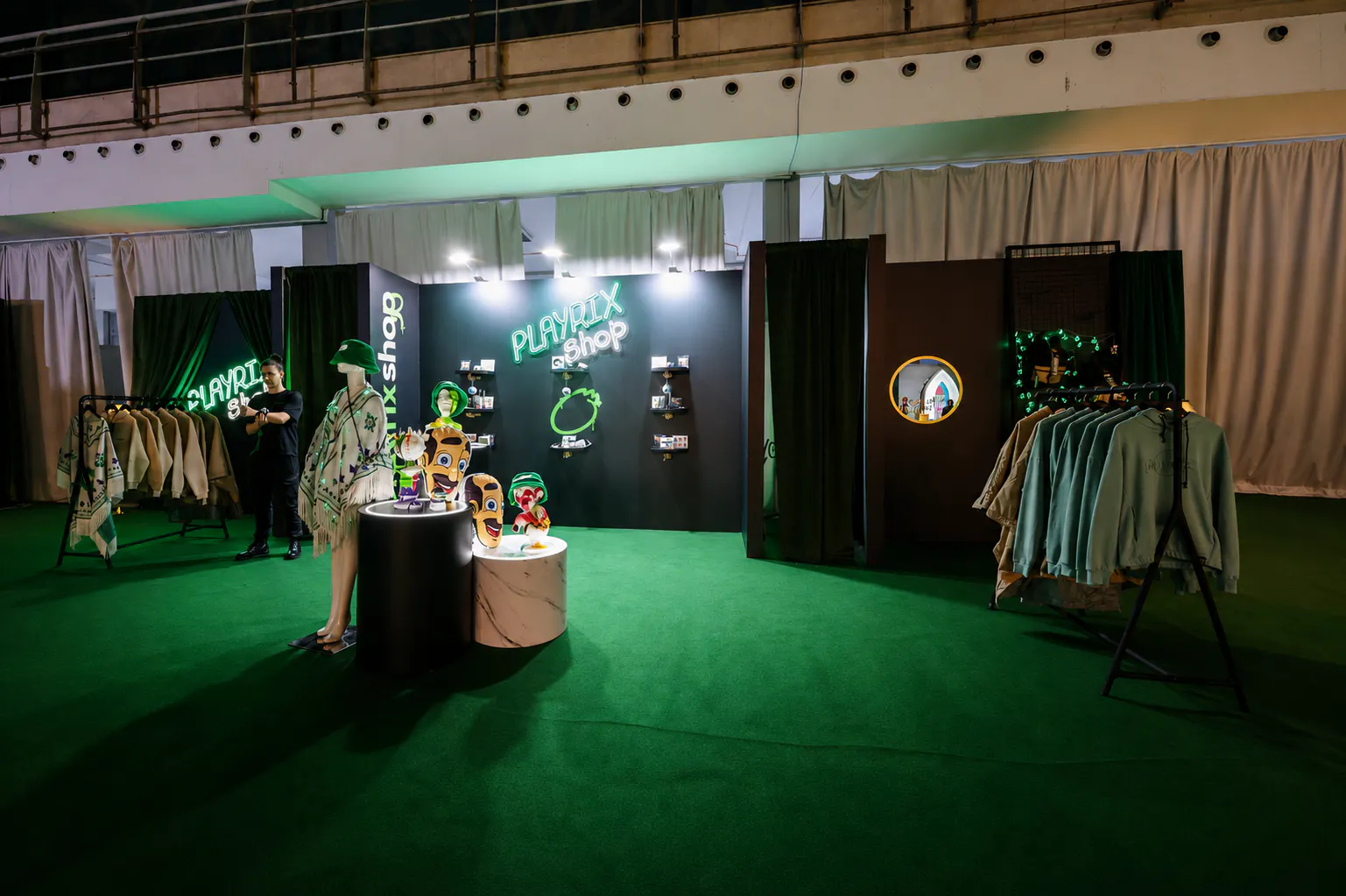 Wide view of the Playrix Shop pop-up with hoodies, ponchos and character busts on green carpet