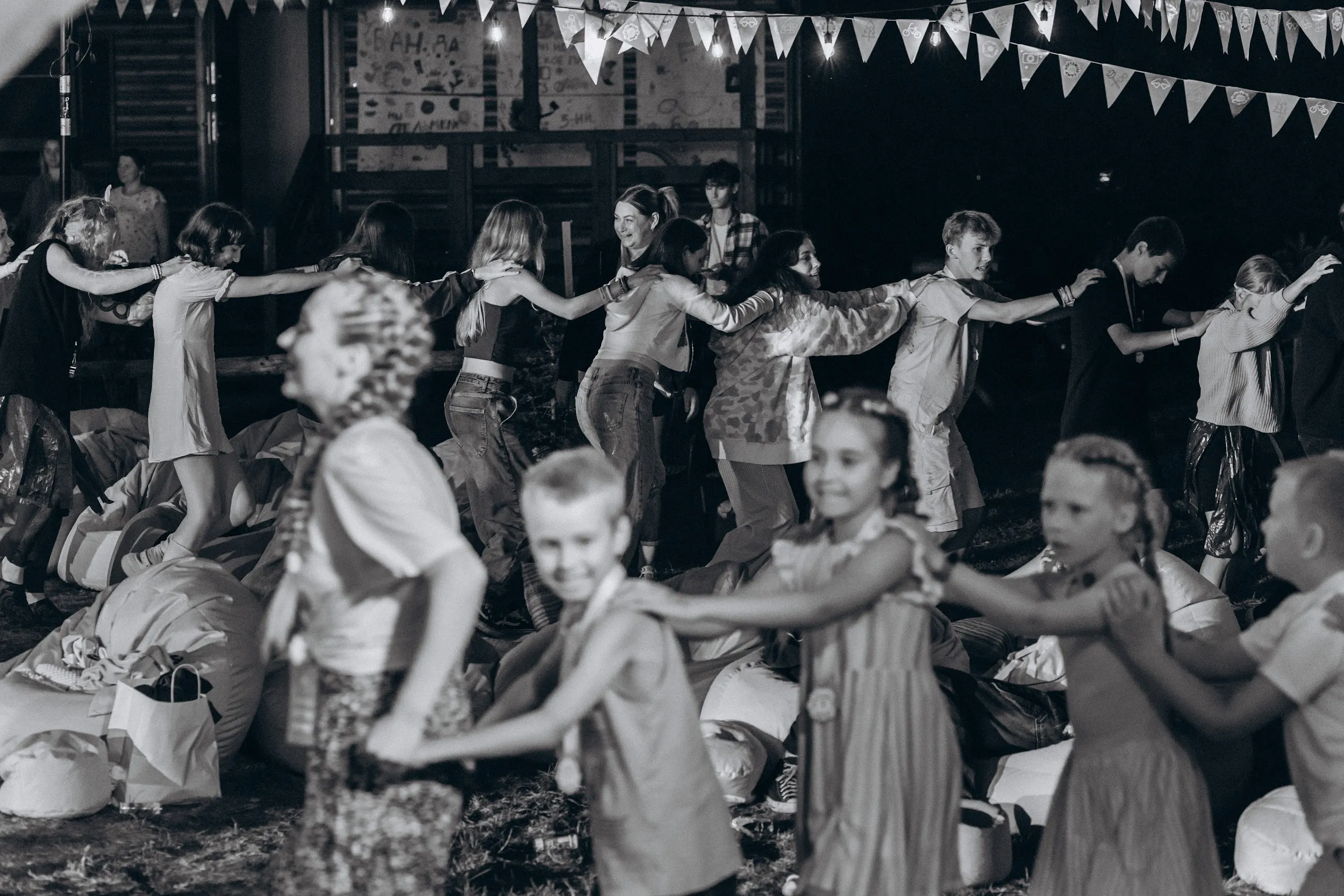Children and counsellors dancing in a conga line at an evening party under bunting lights