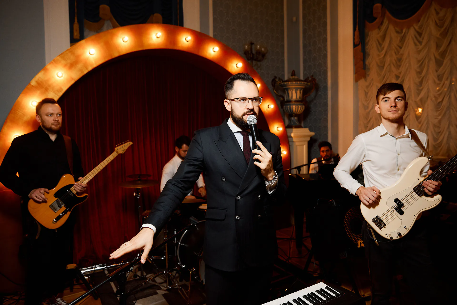 Host with a microphone and a live band on stage under an illuminated marquee-light arch at the Masterday evening gala