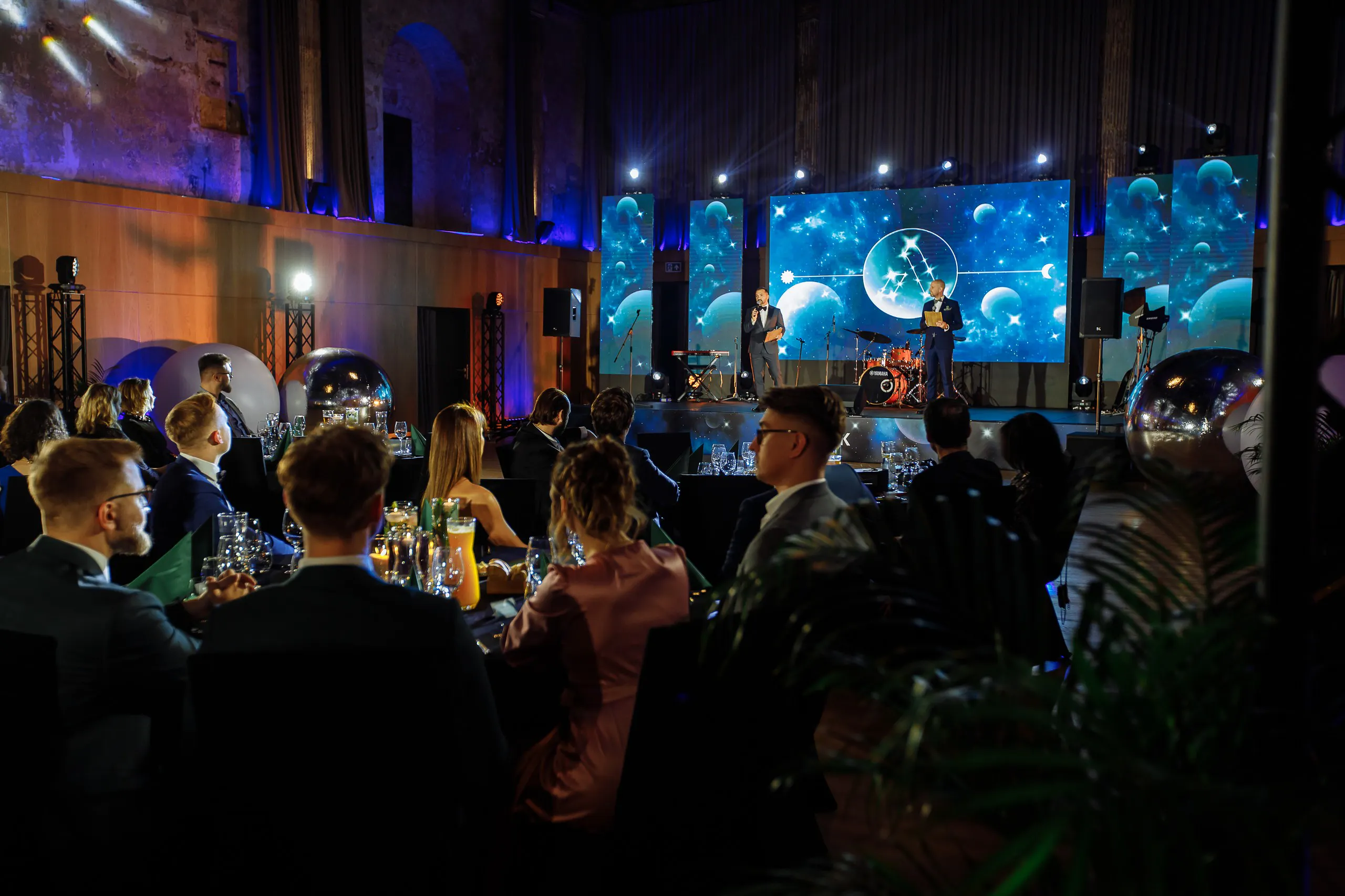 Hosts on stage in front of LED screens with constellation visuals as guests watch the Astek Constellation gala