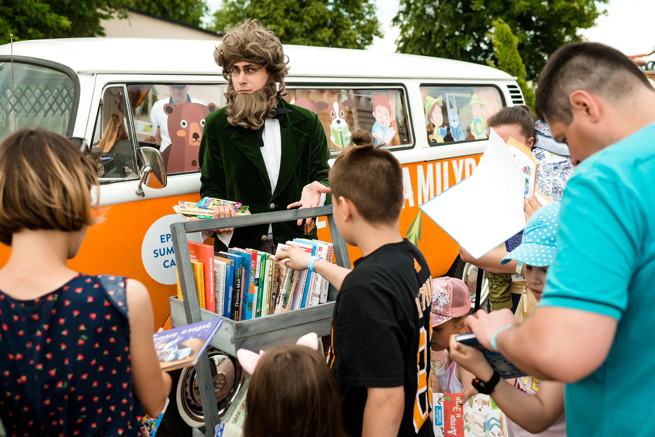 Costumed storyteller presenting children's books from a book cart next to the EPAM Family Day bus