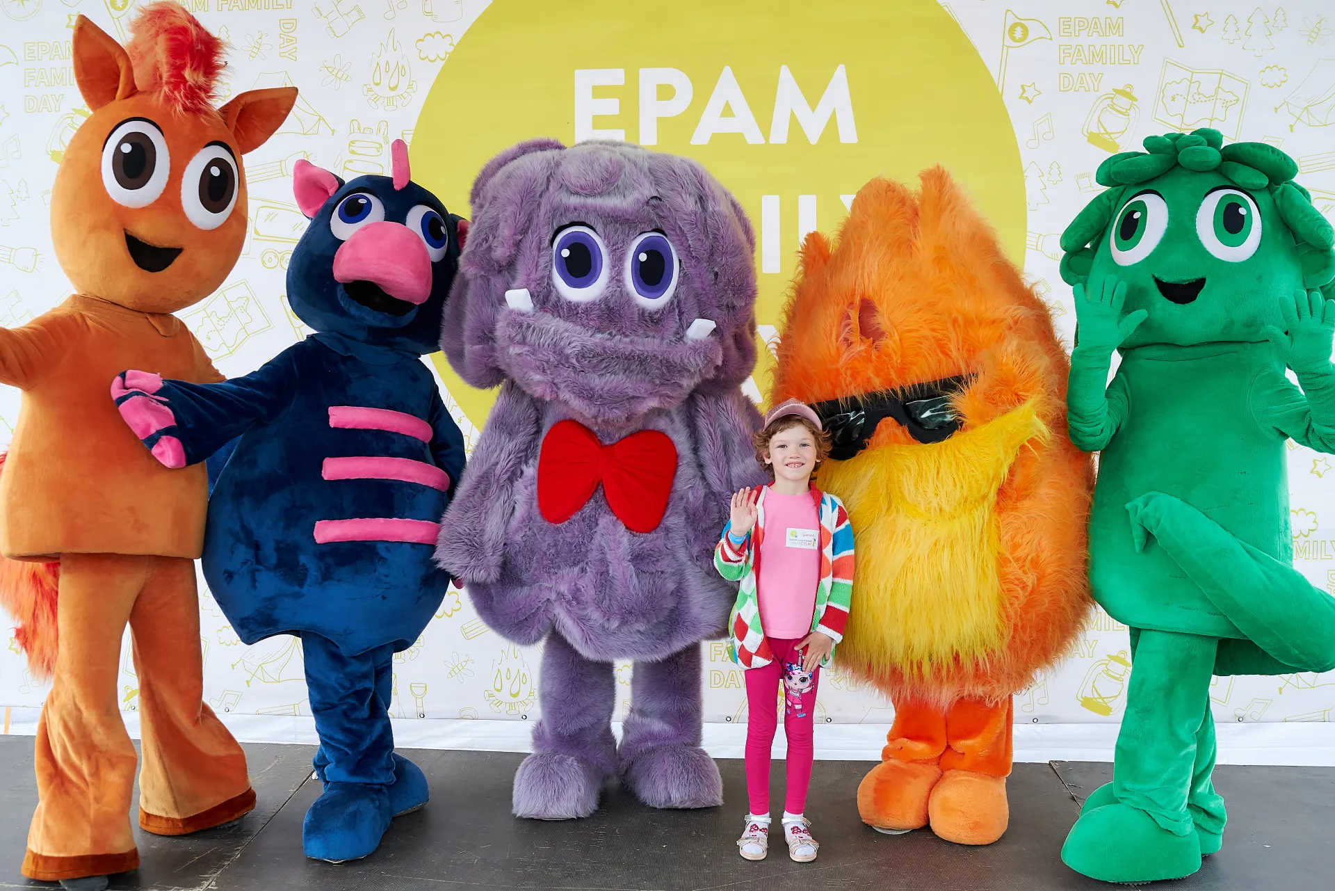 Girl posing with five colorful costumed mascots in front of the EPAM Family Day backdrop