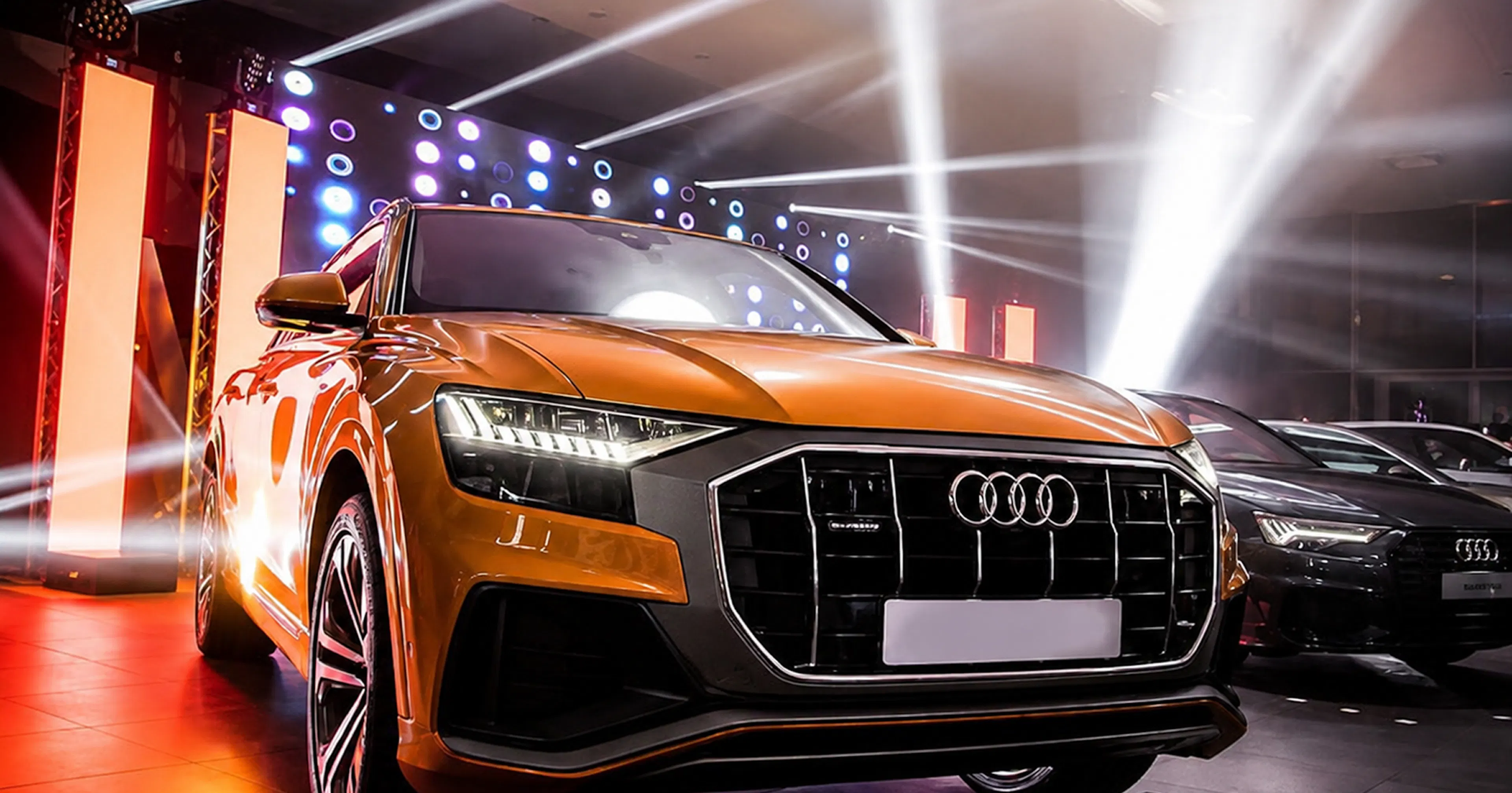 Pomarańczowe Audi Q8 na tle oświetlenia scenicznego i białych snopów światła, obok Audi A6 na premierze Art of Perfection