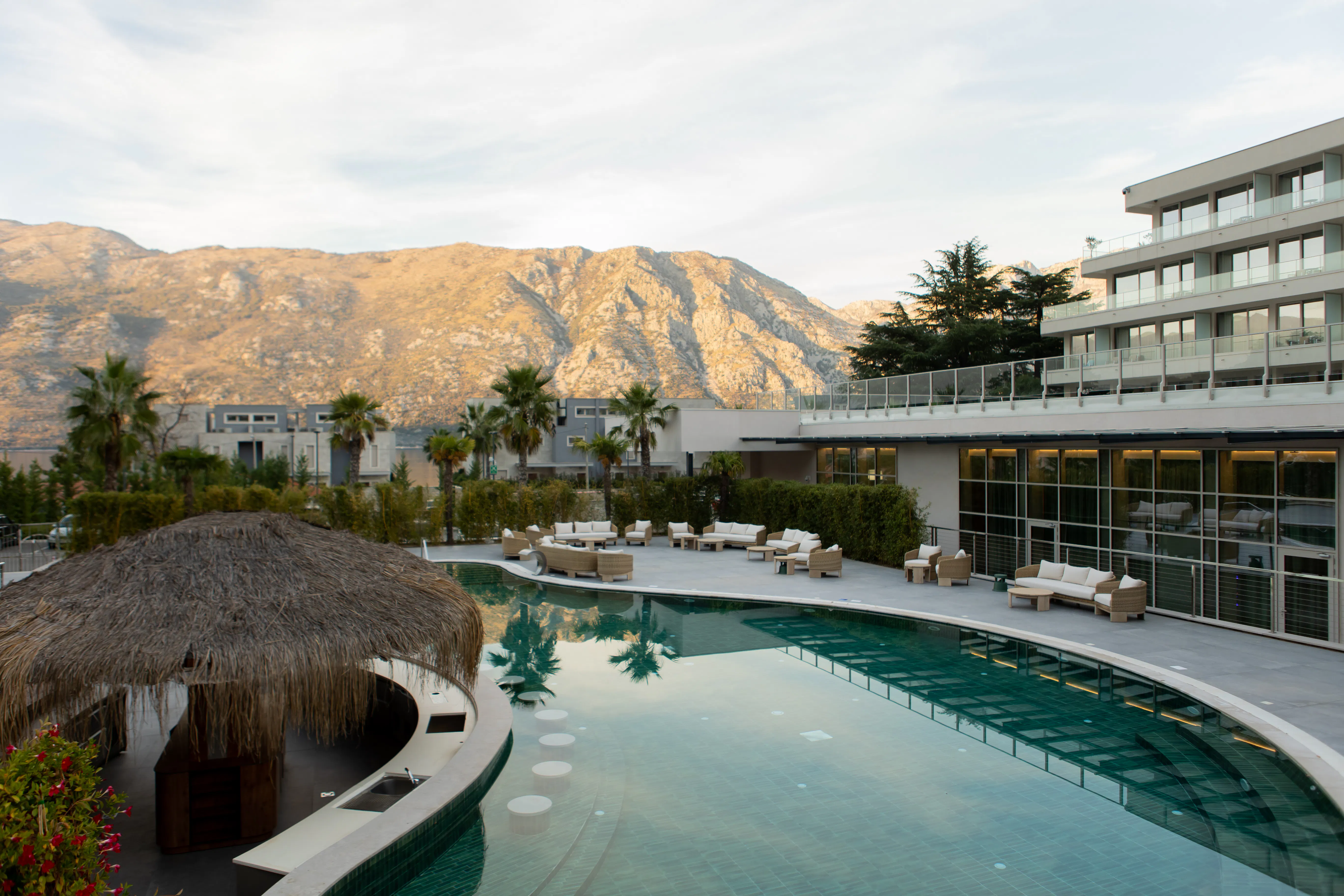 Hotel pool terrace with lounge seating and a thatched pool bar beneath the mountains of the Bay of Kotor
