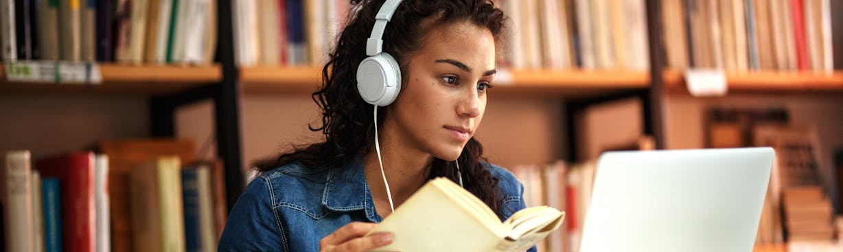 Female in library wearing headphones, holding a book and looking at a laptop screen