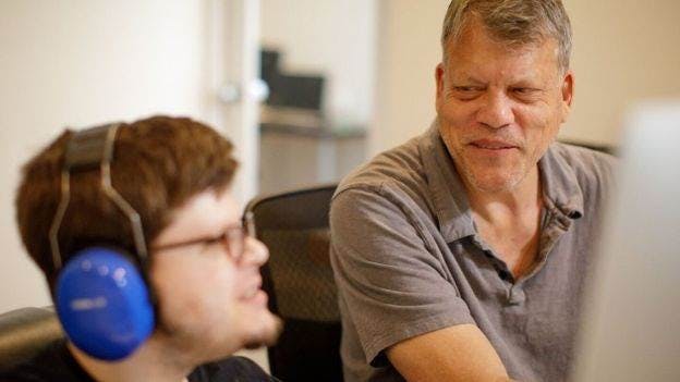 Two auticon employees working at a computer, one wearing noise cancelling headphones