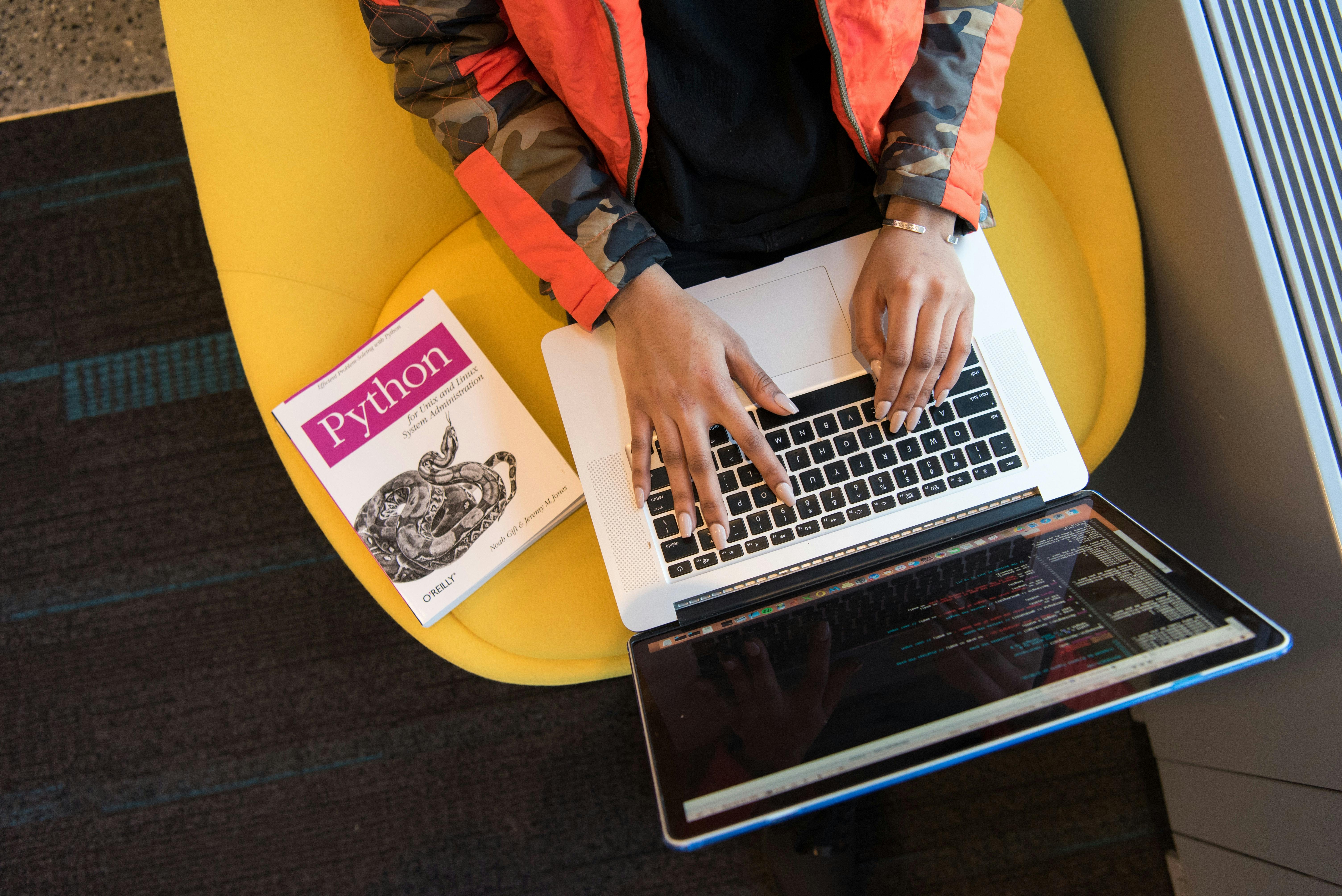 a girl sitting on a yellow sofa, typing codes on the screen with a Python book