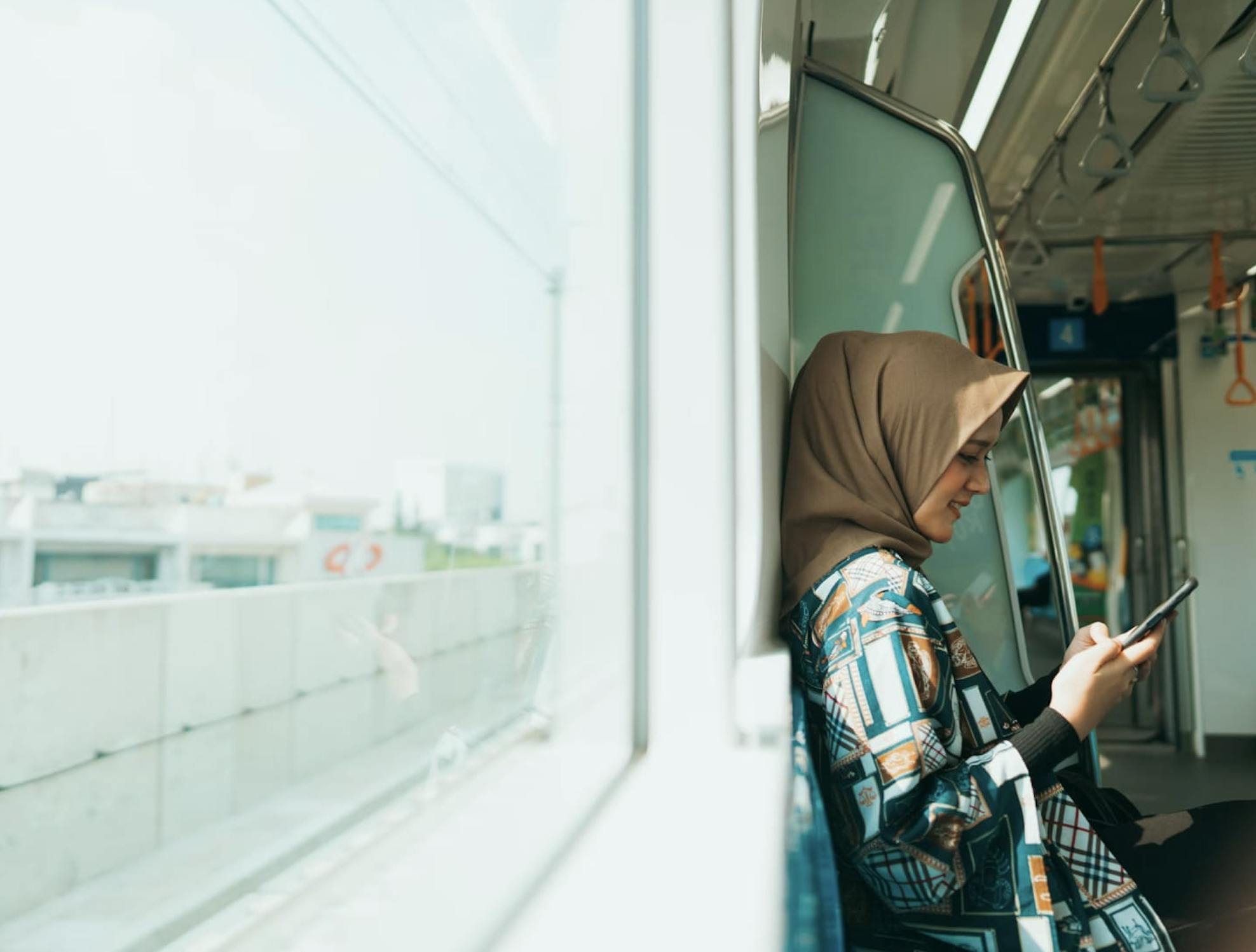 Woman looks at her mobile phone while riding a train, with a large window showing a city view next to her. 