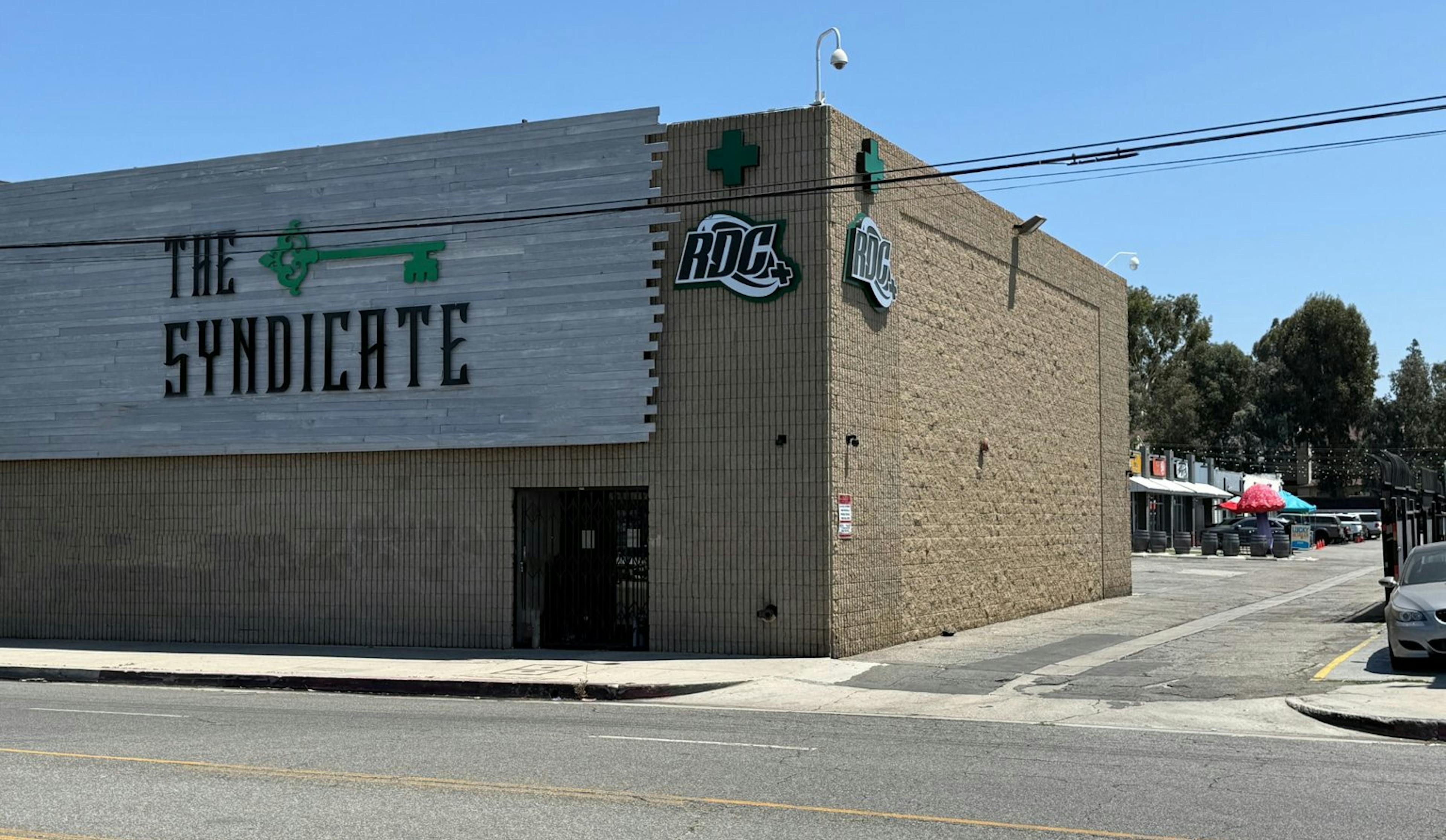 Side view of The Syndicate dispensary exterior, featuring bold branding on gray wood paneling, RDC signage, beige brick walls, and neighboring retail storefronts under a clear blue sky.