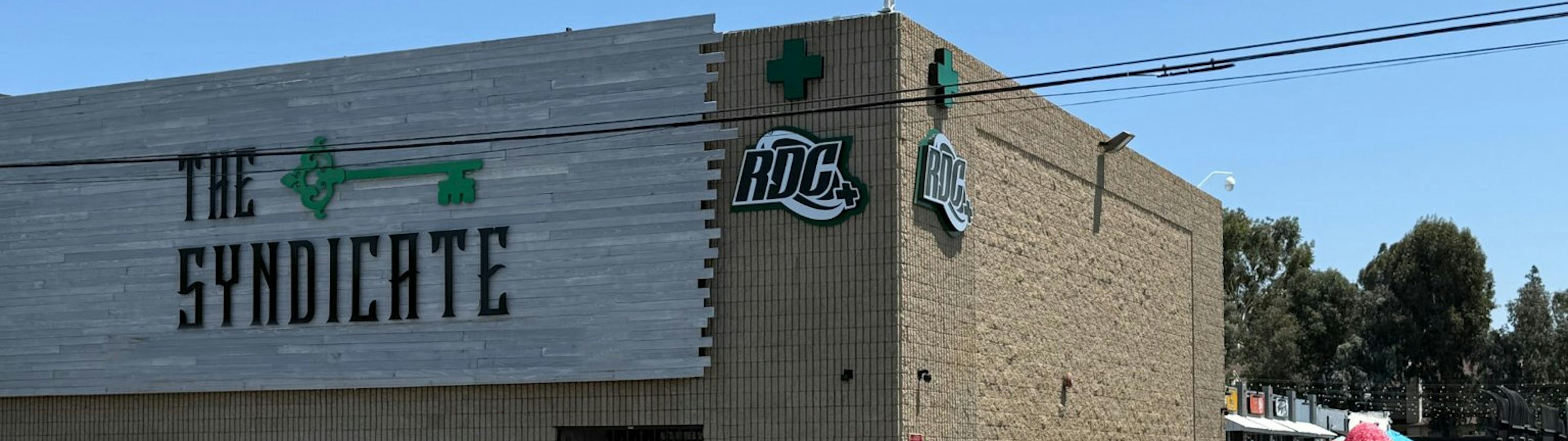 Side view of The Syndicate dispensary exterior, featuring bold branding on gray wood paneling, RDC signage, beige brick walls, and neighboring retail storefronts under a clear blue sky.