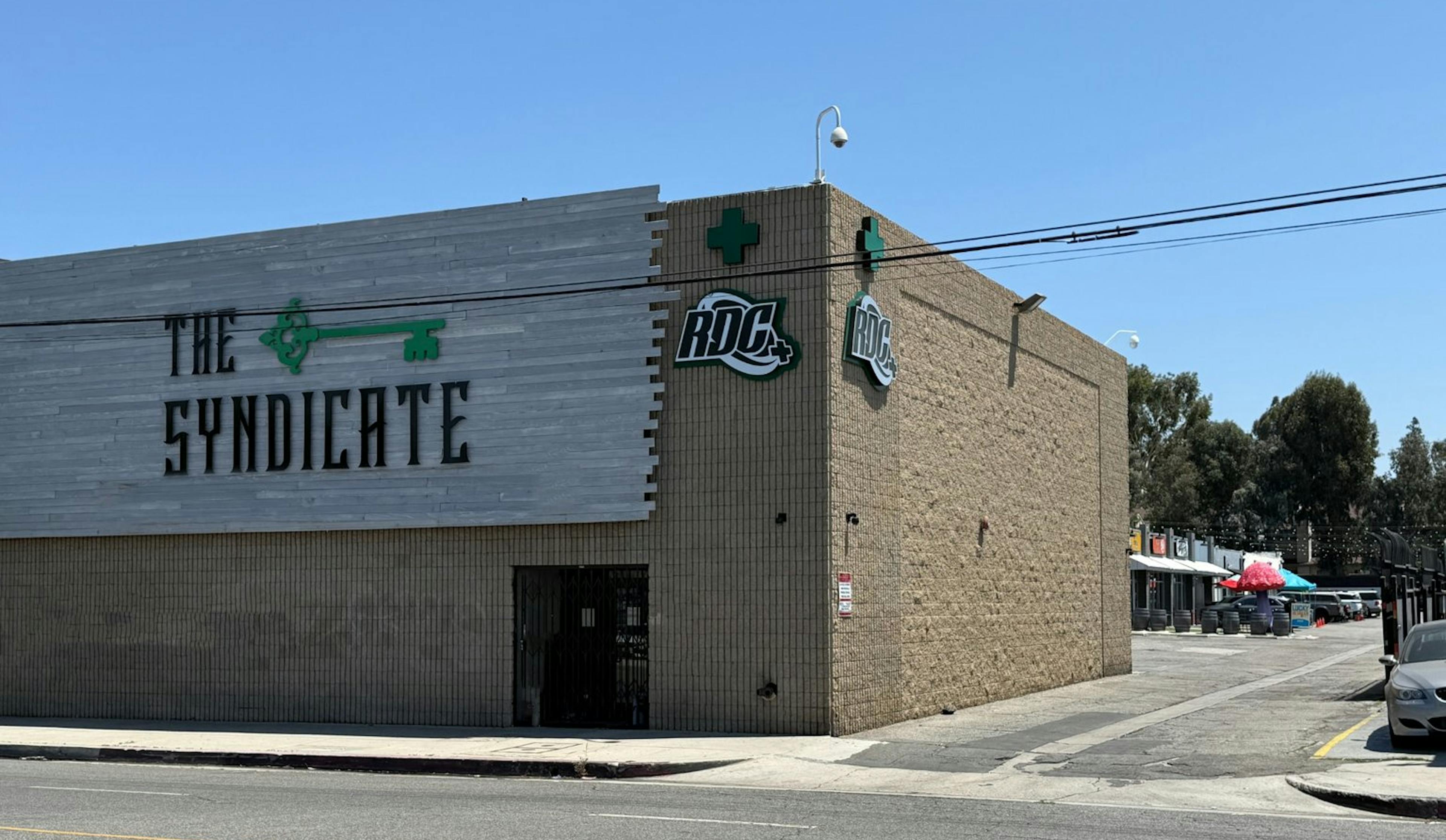 Side view of The Syndicate dispensary exterior, featuring bold branding on gray wood paneling, RDC signage, beige brick walls, and neighboring retail storefronts under a clear blue sky.