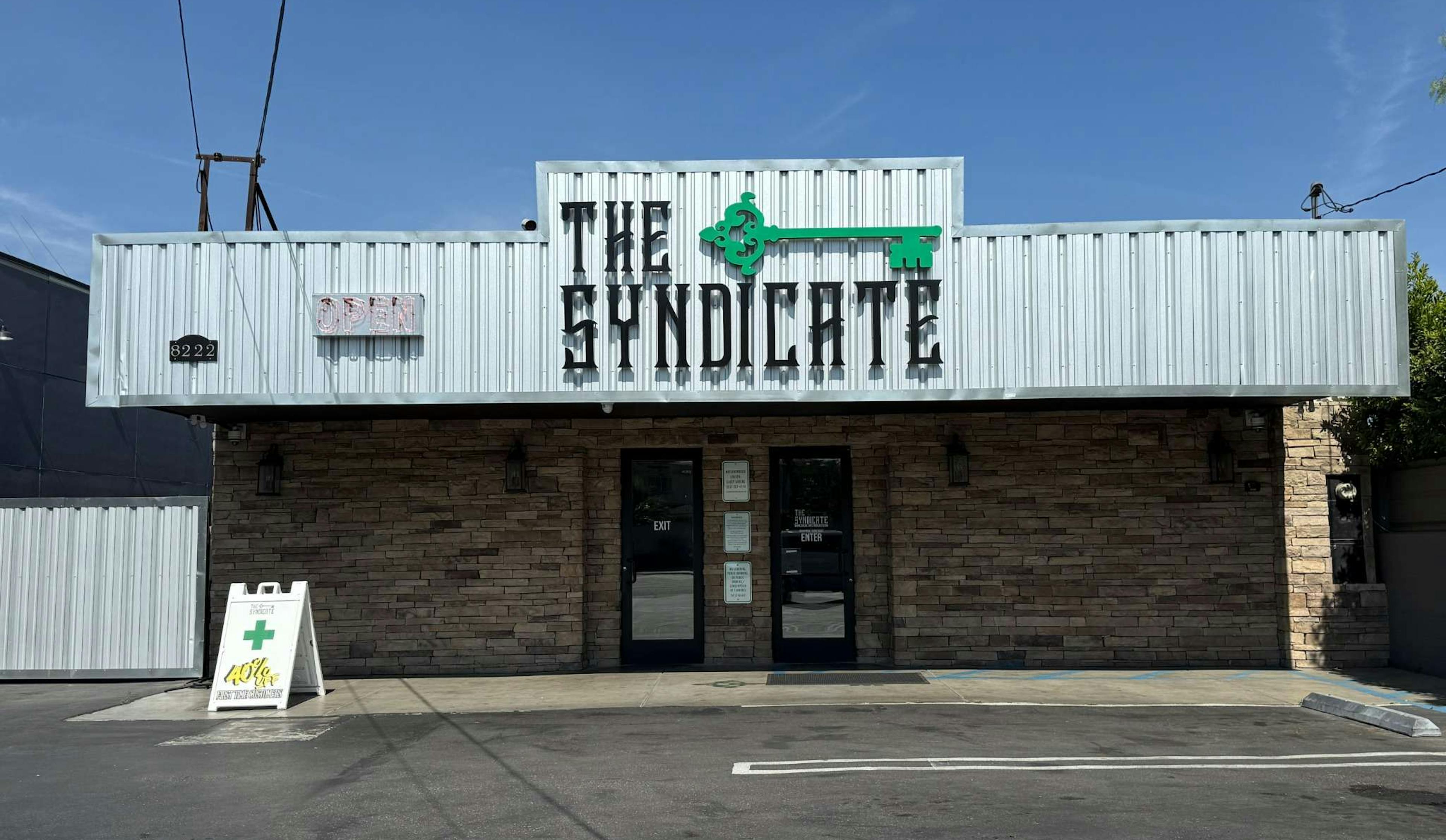 Front view of The Syndicate storefront at 8222, featuring a stone facade, metal siding, a green key logo, and an A-frame sign advertising a 40% discount.