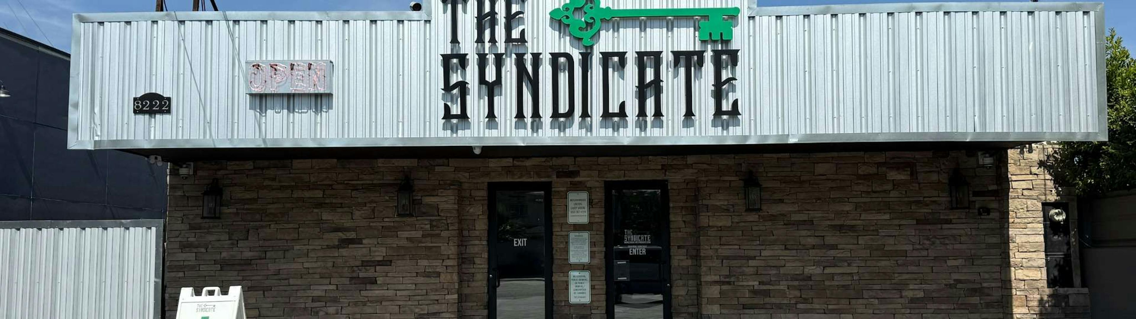 Front view of The Syndicate storefront at 8222, featuring a stone facade, metal siding, a green key logo, and an A-frame sign advertising a 40% discount.